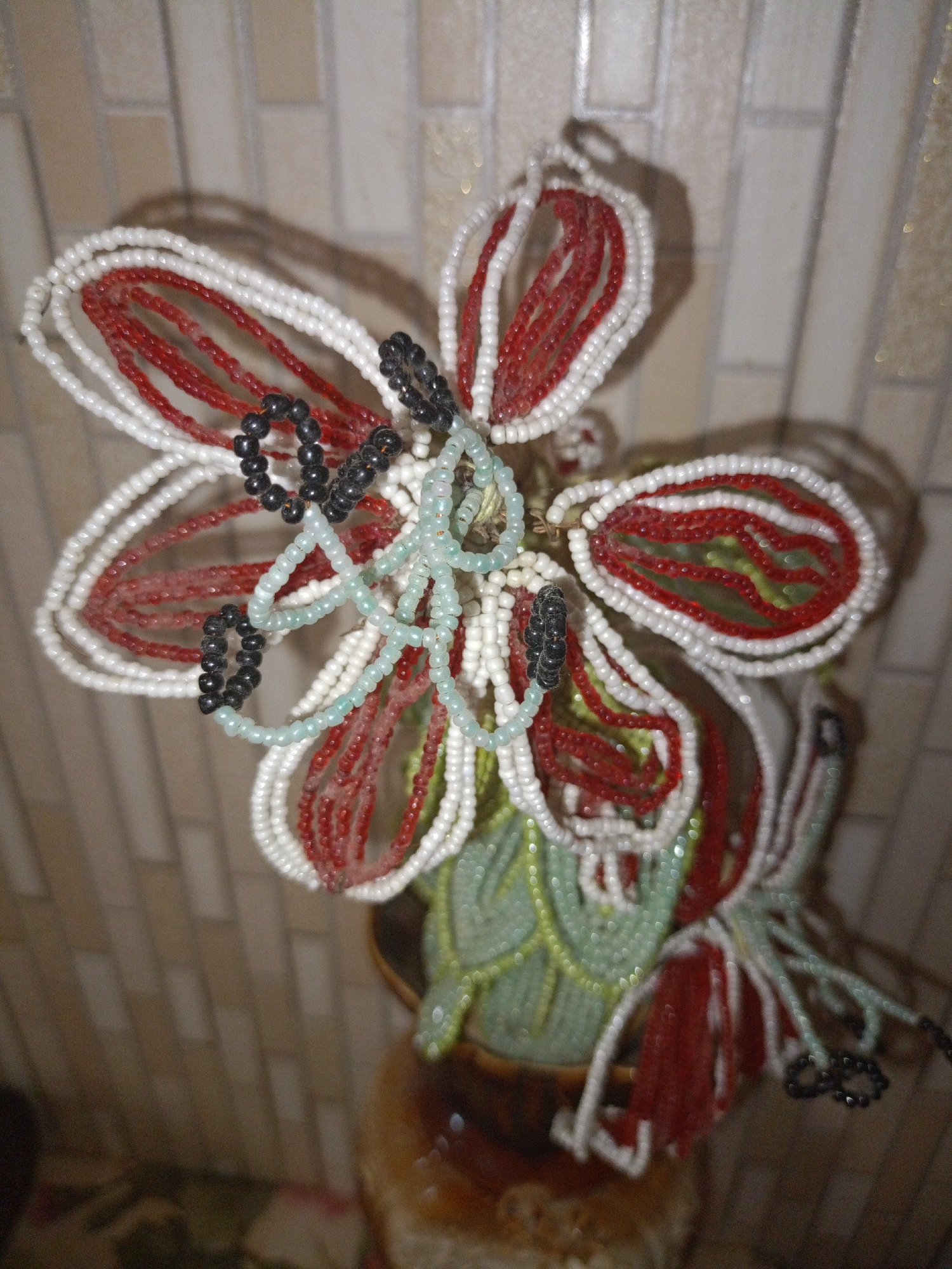 A beaded floral arrangement is displayed in a pot. The arrangement features white, red, and light green beads, with some black beads forming loops. The beads are meticulously crafted to resemble petals and leaves. The background consists of a tiled wall with a light beige color and a grid pattern. The lighting in the image is warm, casting soft shadows and highlighting the textures of the beads and the wall. The location of the object is in the city of Mytishchi, Russia.