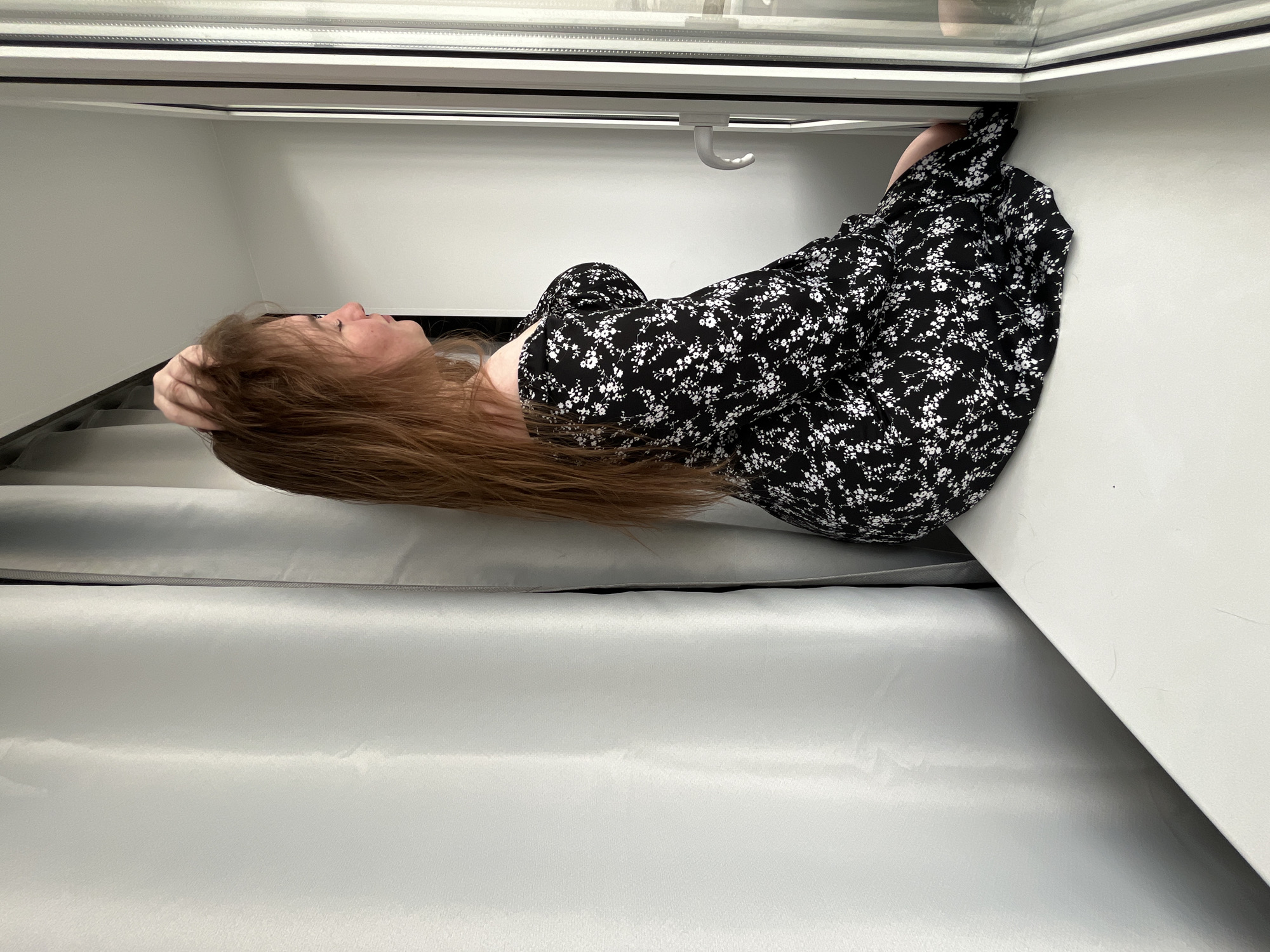 A young woman with reddish-brown hair is positioned near a window in what appears to be an interior space. She is wearing a black dress with a white floral pattern. Her left hand is raised to her head. The background shows a white wall and a window frame with a metal bar running across the top. Additionally, there's a hanging hook-like object. Below her, there are grey curtains. There is no clear evidence to definitively state the location is Chaikovsky, Russia, or even that it is indoors.