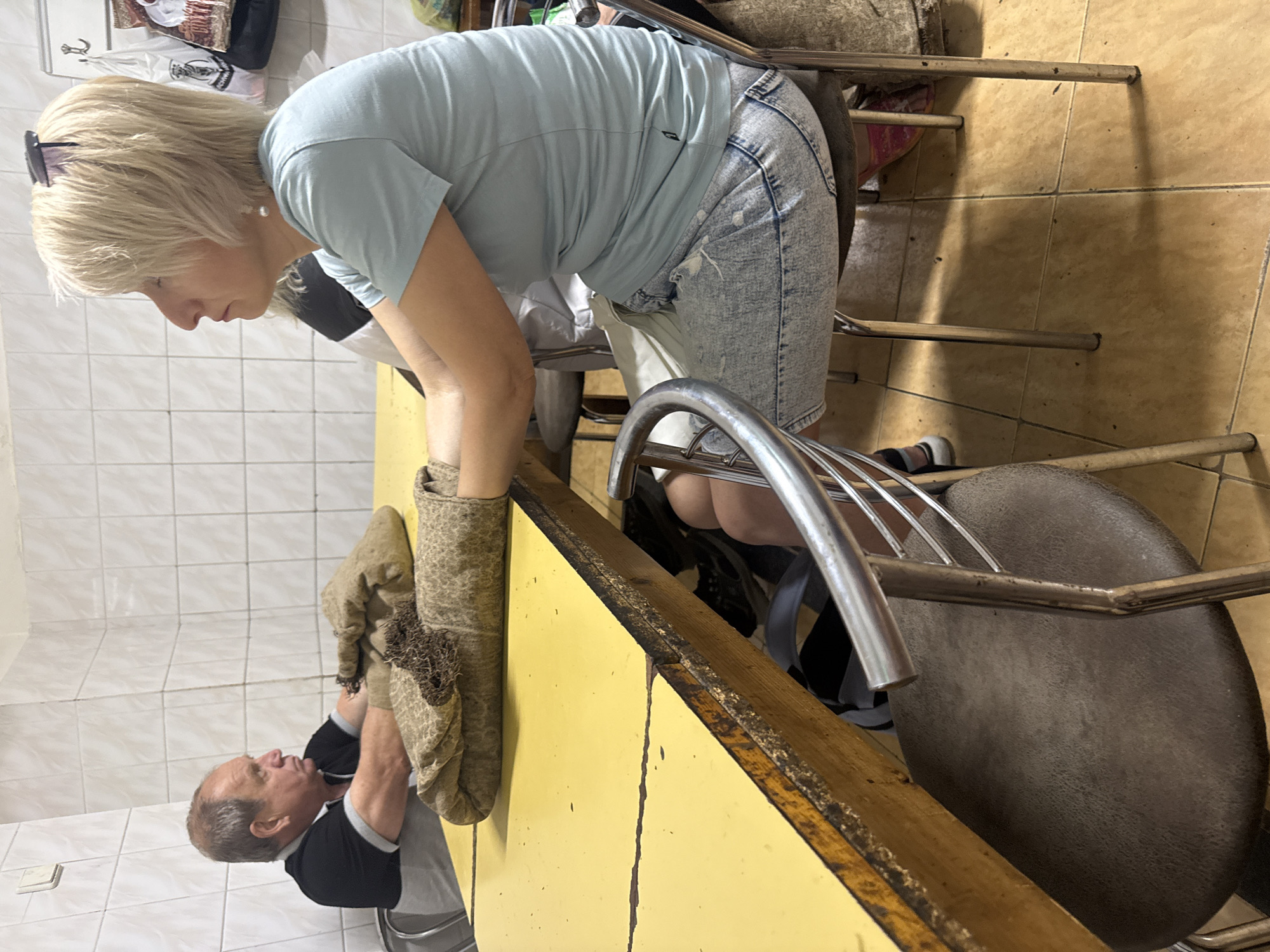 A woman with short blonde hair wearing a light blue t-shirt and acid-wash denim shorts is bending over a yellow counter. Across the counter, a man in a black and gray shirt holds up a pair of brown mittens. The scene appears to be indoors, with tiled walls, possibly in Syniak, Ukraine. A metal chair with a brown seat is visible, along with sections of other chairs, indicating this might be a kitchen or eating area. The overall lighting is bright.