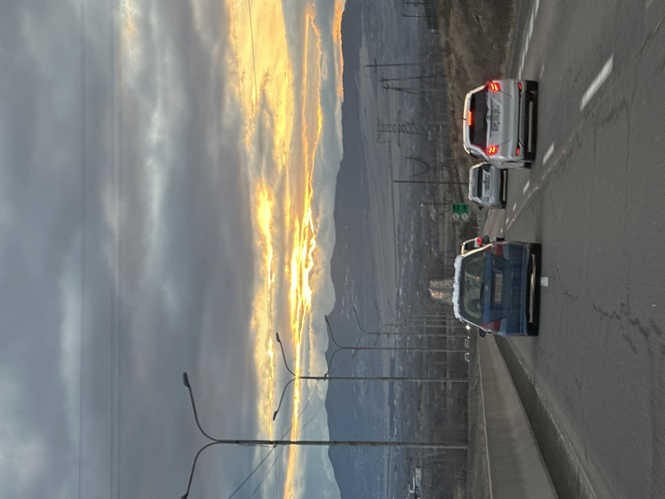 A cloudy sky with streaks of bright orange light flanks a roadway with cars. Three vehicles are visible, traveling on the right side of the road, a blue car in the foreground, followed by two white cars. Power lines and streetlight posts run along the edge of the highway. Hills and a cityscape are visible in the distance under a hazy sky.