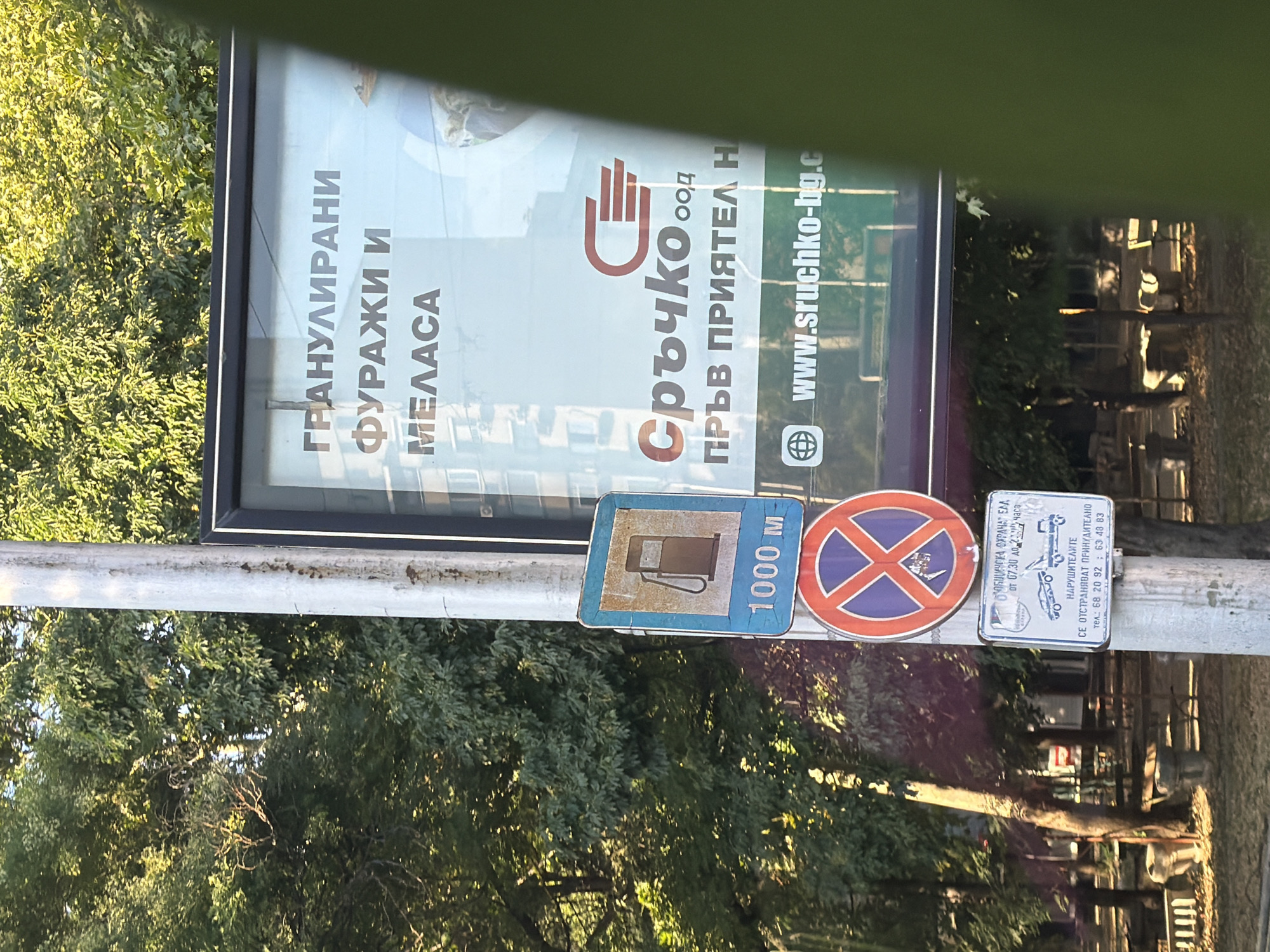 The image shows a street scene with a light pole featuring several signs and a large billboard. The billboard displays text in Cyrillic: "ГРАНУЛИРАНИ ФУРАЖИ И МЕЛАСА" (Granulated Feed and Molasses), and the company name "Сръчко" (Srchko) with the text "пръв приятел н" (first friend n). Below this is a website address, "www.srchko-bg.c".
Attached to the light pole are three signs. The top sign is a blue square with a white border, depicting a gas pump icon in brown. Below the icon, "1000 M" is written in white text. The middle sign is a circular red and blue prohibition sign with a white "X" in the center. The bottom sign is a rectangular white sign with Cyrillic text and illustrations. The text describes services related to towing vehicles and mentions "НАРУШИТЕЛИТЕ" (Offenders) and contact phone numbers: "тел. 68 20 92 | 63 48 83". The illustrations show a tow truck and a car being towed.
The environment appears to be an outdoor urban setting with trees providing shade. The background shows buildings and what appear to be park benches. The lighting suggests it is daytime, with sunlight filtering through the leaves.
Based on the visible information, particularly the company name "Srchko" and the Cyrillic script, the location is likely Bulgaria. The billboard's advertising content and the street signage are typical of an urban or suburban environment. Without more specific landmarks or context, pinpointing the exact location within Plovdiv, Bulgaria, is not possible from this image alone. However, the presence of such advertising and street signs is consistent with an urban area in Bulgaria.