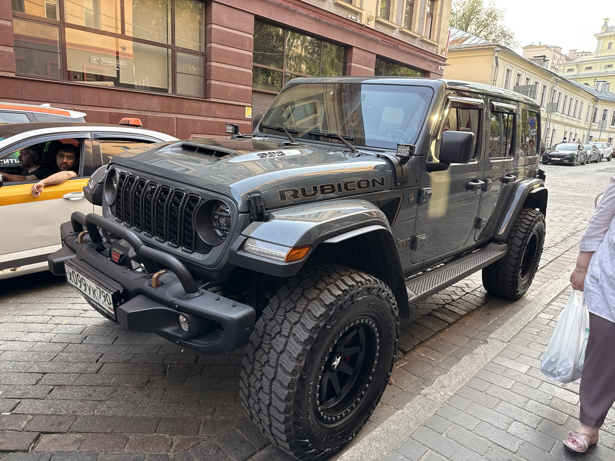 The image depicts a gray Jeep Rubicon parked on a cobblestone street in Moscow, Russia. The vehicle is in the foreground, angled toward the viewer. The Russian license plate "X099YK790 RUS" is visible on the front bumper. The exterior of the car appears to be in good condition. In the background, a yellow and white taxi is partially visible with two male figures inside. A woman is also visible on the right side of the frame with a shopping bag in hand. The architecture suggests a European city setting, with buildings having multiple windows.