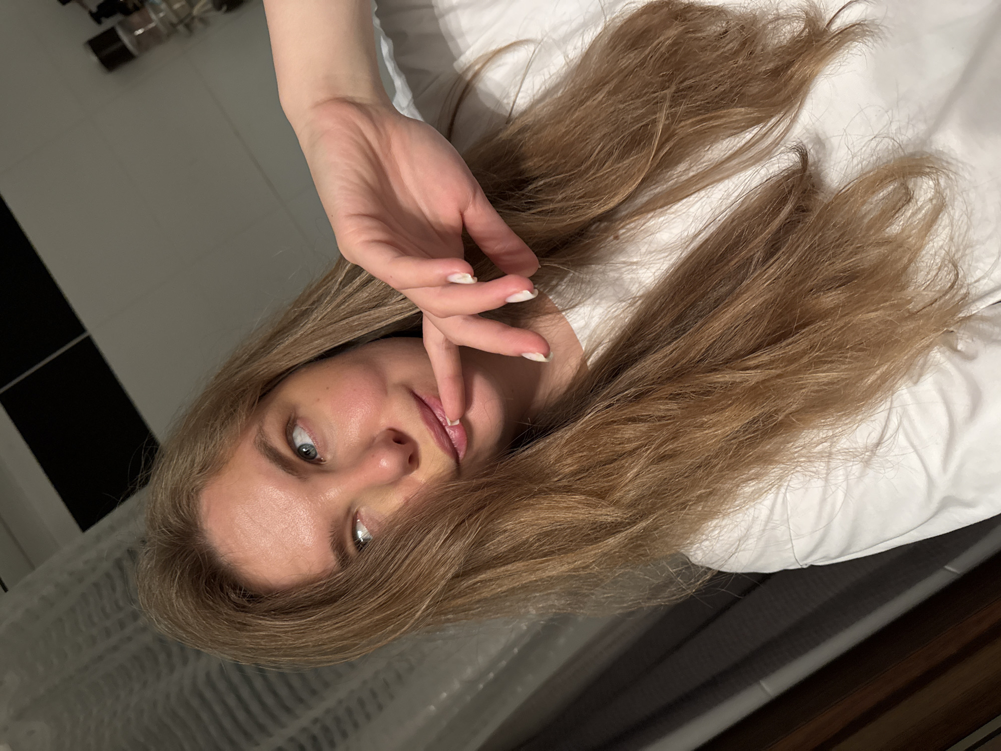 A young woman with long, light brown hair and blue eyes is shown in a close-up, selfie-style shot. She is wearing a white t-shirt and her right hand is touching her lips. Her gaze is directed upwards and to her left. The background appears to be an indoor setting with a tiled wall, a shower curtain, and some shelves with bottles, possibly a bathroom. The lighting is dim. The image was taken in a residential building in Vladimir, Russia.