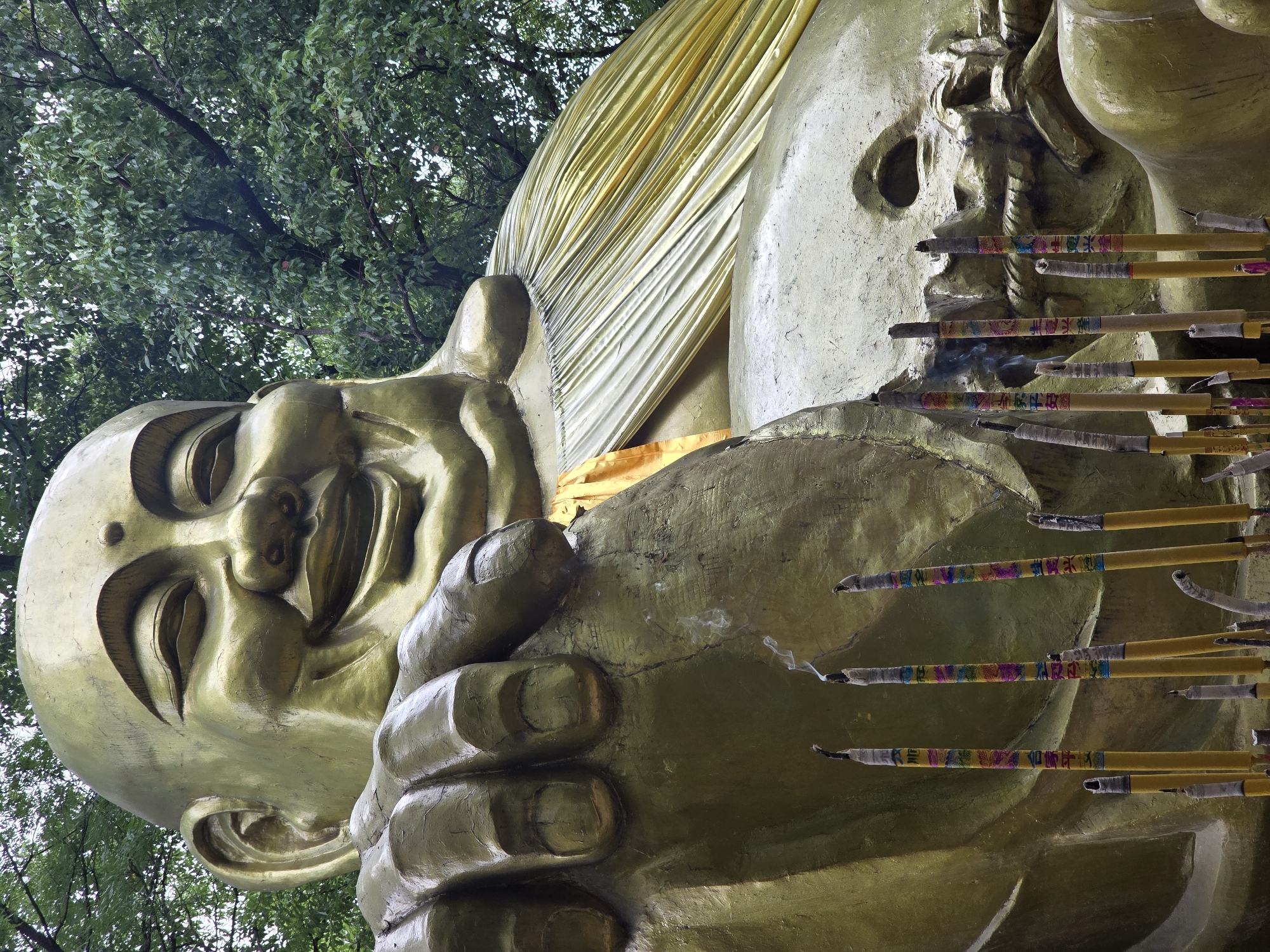 This is a medium shot, eye-level, vertical photograph of a large, golden statue of the Budai, also known as the "Laughing Buddha," set against a backdrop of lush green foliage. The statue is depicted with a rounded, bald head, a broad smile, and closed eyes. Its right hand is resting on its chest, while its left arm is bent and partially obscured. A yellow sash is draped over the statue's shoulder and flows down its body. In the foreground, a cluster of lit incense sticks smolders, with wisps of smoke rising. The incense sticks are cylindrical and appear to be decorated with colorful patterns and Chinese characters. The statue is located in Xihu District, China, likely within a temple or garden. The overall lighting suggests an overcast or shaded environment, contributing to the subdued yet vibrant tones of the image.
