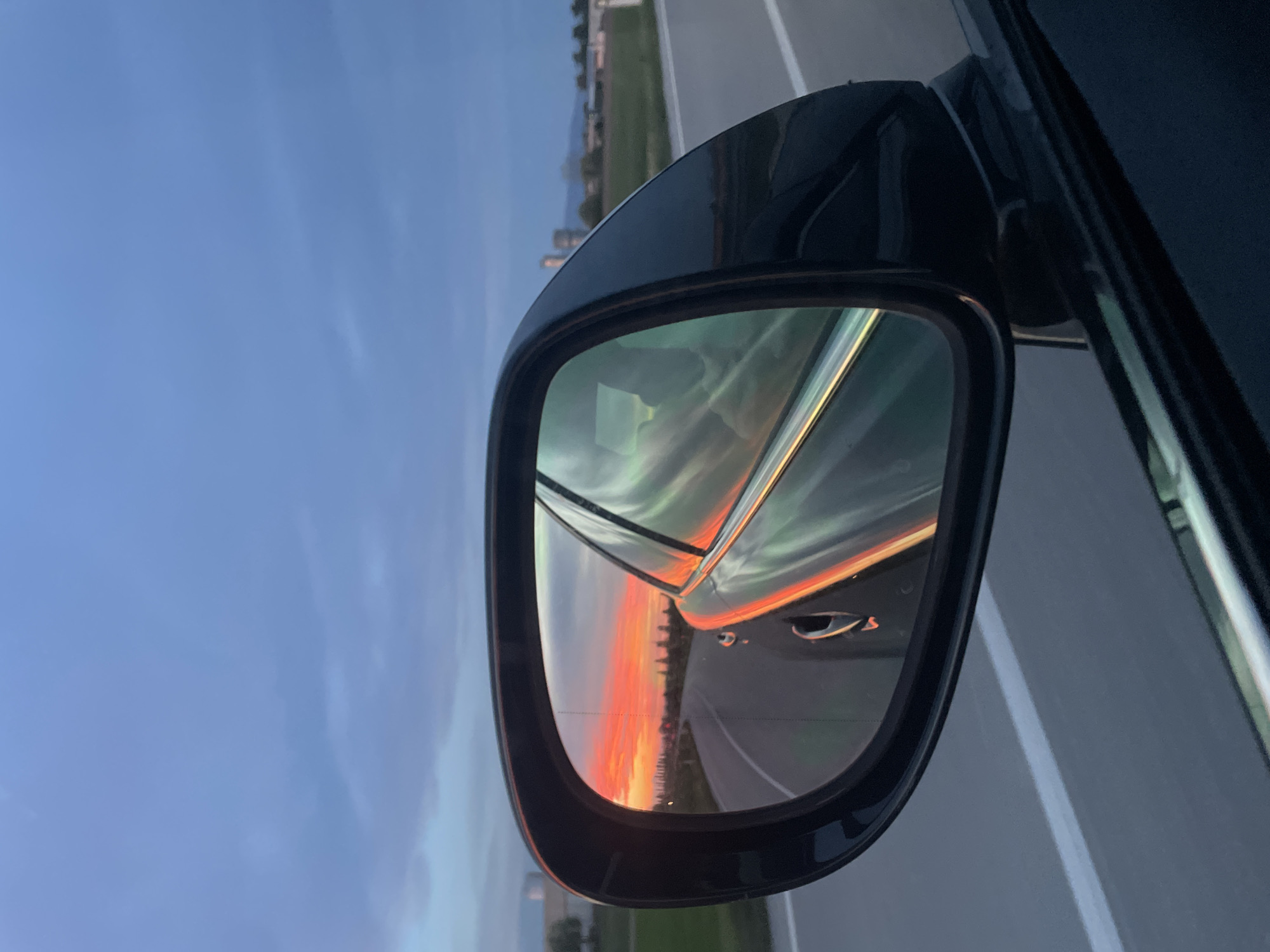 A photograph depicts a car's side mirror reflecting a vibrant sunset. The mirror's reflection shows a strip of orange and red hues along the horizon, transitioning to softer green and blue tones above. The road surface and a portion of the car's door are also visible within the mirror. Outside the mirror, a segment of the highway with white lane markings is seen, with buildings and some greenery in the distance. The sky transitions from blue to a lighter hue. The location is likely not San Lorenzo, Italy, but rather a highway near a city. The image does not provide explicit information to pinpoint the exact location.