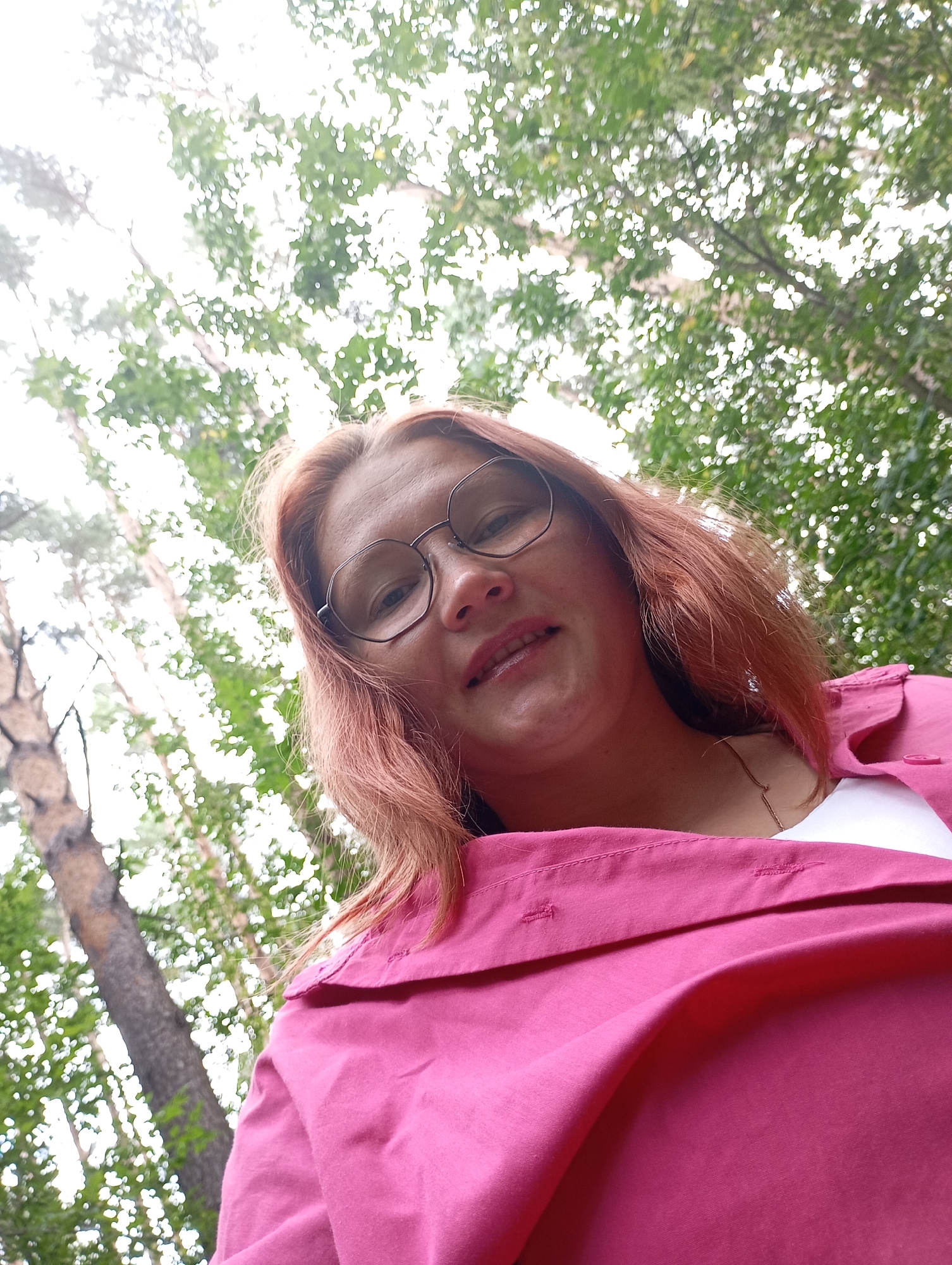 A low-angle, medium shot captures a woman from the chest up, outdoors amidst a forest. She has fair skin, light brown hair, and is wearing round, metal-rimmed glasses. Her mouth is slightly open, revealing her teeth, and she has a hint of a smile. She is dressed in a bright pink collared shirt over a white undershirt, with a thin gold necklace visible. The background is dominated by the dappled sunlight filtering through the leaves of tall trees, with a thick tree trunk visible to the left. The scene appears to be a casual outdoor portrait.