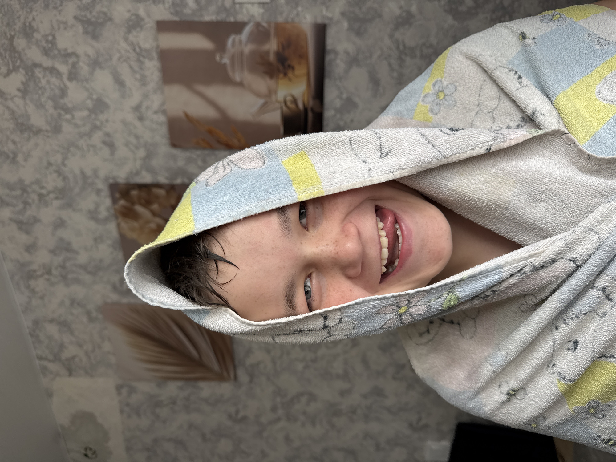 This image, taken in Chaikovsky, Russia, features a young individual, likely a child or teenager, captured in a joyful and playful moment. The person is wrapped in a hooded towel, suggesting they have just had a bath or shower, as their short, dark hair appears wet. They are looking directly at the camera with a wide, open-mouthed smile, playfully sticking out their tongue. Freckles are visible across their nose and cheeks, and they have light-colored eyes.
The hooded towel is adorned with a pattern that includes sections of white, light blue, and yellow, with faint floral or abstract designs. The background is a wall covered in a textured, grayish wallpaper. On this wall, two framed pictures are visible. The upper frame shows a transparent teapot, possibly containing tea or herbs. Below it, another frame displays a botanical image, perhaps a dried palm leaf or stalks of wheat, adding a natural touch to the indoor setting. The image is rotated 90 degrees counter-clockwise, making the subject appear to be looking upwards from the right side of the frame. There is no visible text in the image.