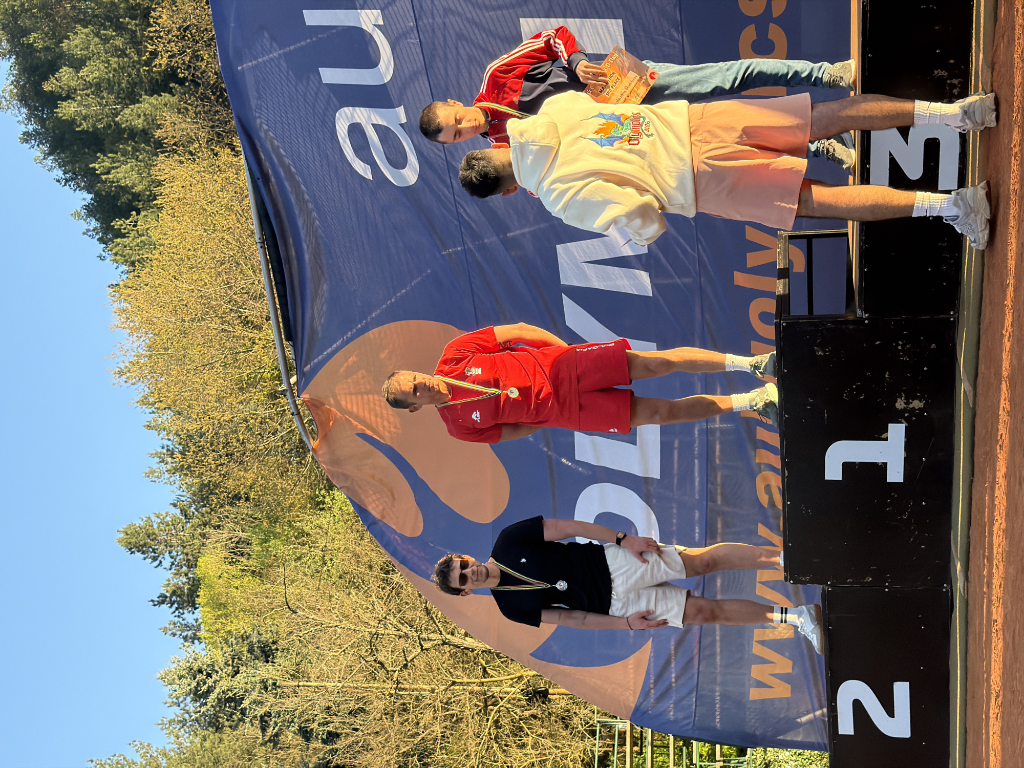 The image shows a tennis tournament medal ceremony. Three athletes stand on a black podium with the numbers "2", "1", and "3", indicating their placement. From left to right, the athletes are wearing: a black t-shirt with white shorts and sunglasses; a red t-shirt with red shorts; a white hoodie with pink shorts; and a red tracksuit. All of them are wearing medals. A large blue banner with orange and white writing is in the background. The environment appears to be outdoors on a sunny day, with trees visible behind the banner and a clay-like tennis court surface in the foreground. The banner shows the words "OLYM" "WWW OLY" and "CS", and "Olympics 2026" as well as "GO D-Day", "MAN HON", "3RD PLACE", and "notantin Dimibrov"