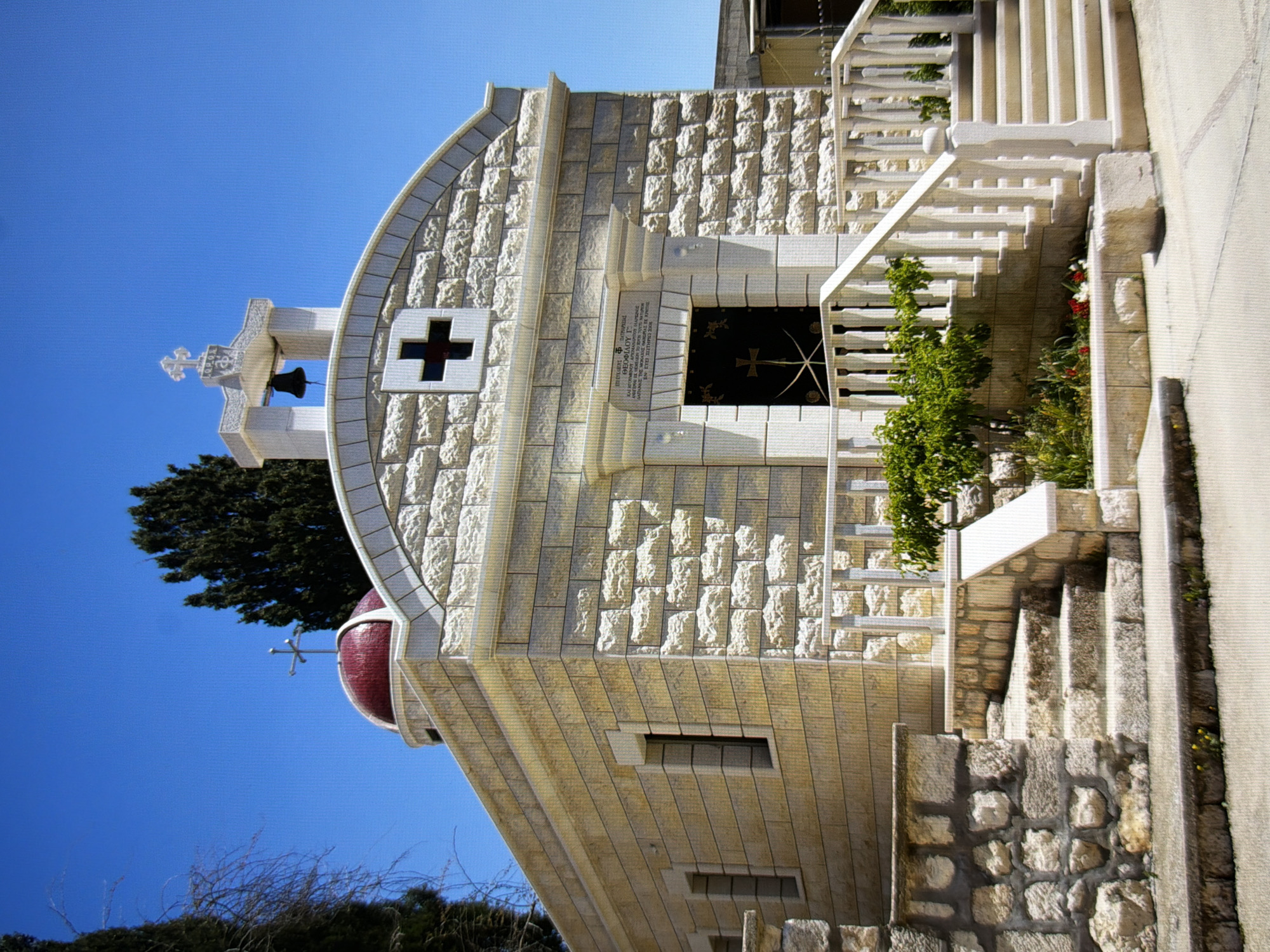 The image shows a small, light-colored stone building, possibly a chapel or church, with a red-domed roof and a cross on top. The facade features decorative stonework, a dark-colored door with a cross and swords emblem, and an arched opening above the door with Greek inscriptions. A bell is visible in a bell tower structure above the main entrance. The building is approached by a set of stone steps with a white balustrade, and there are some green plants and flowers in front of the steps. The building is situated outdoors under a clear blue sky. There are no people visible in the image. The architectural style appears to be Byzantine or Orthodox Christian. The image is a photograph of the building's exterior. There is no indication of the location being Kyiv, Ukraine.