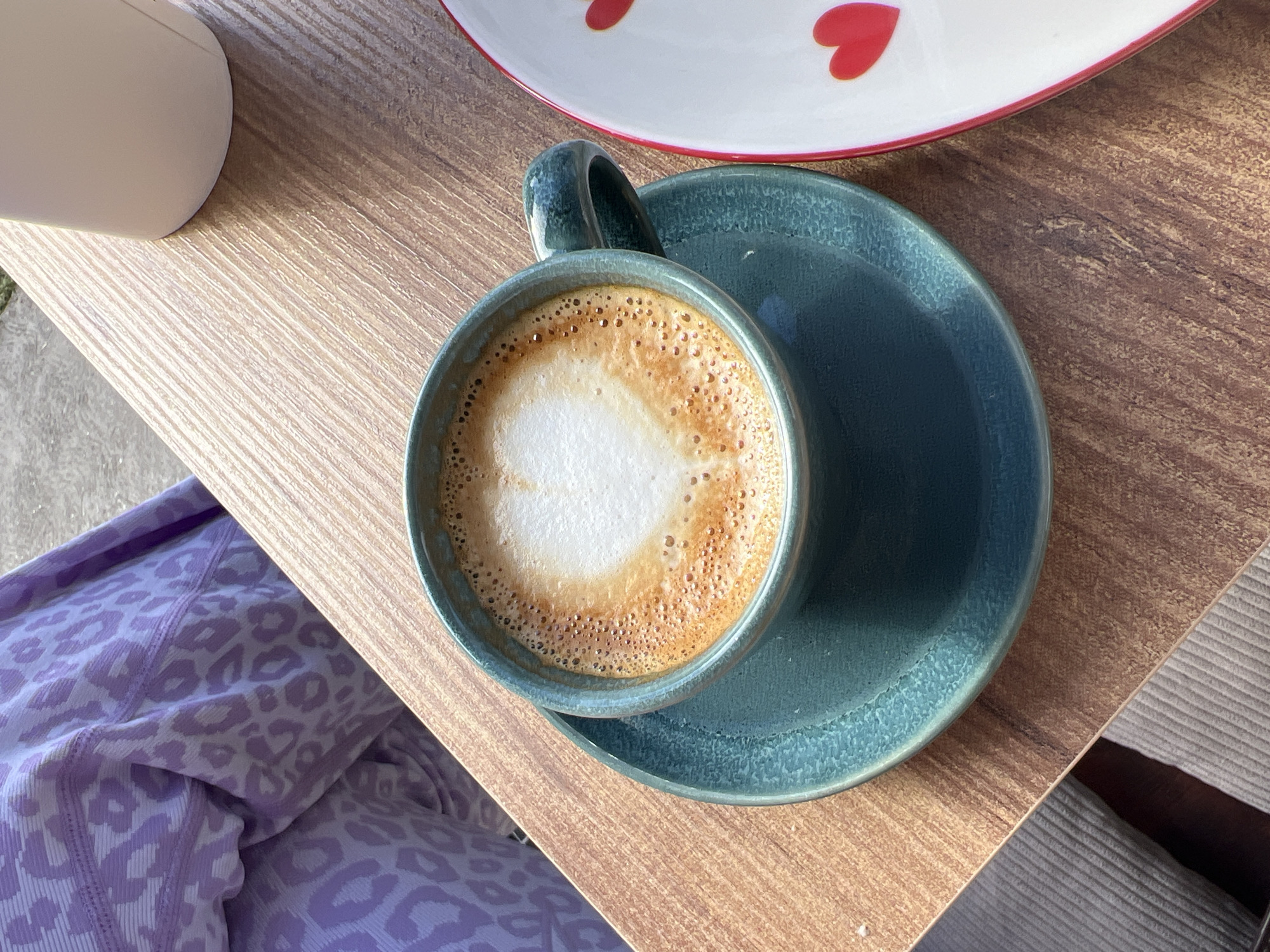 A top-down shot shows a cup of cappuccino in a dark teal ceramic mug and saucer, resting on a wooden table. Beside it is a white ceramic plate with red heart decorations along the rim. A white mug is partially visible on the left side of the image. The bottom left corner shows a section of fabric with a light purple leopard print pattern. A portion of striped fabric is seen at the bottom right corner. The table surface appears to be textured with a linear pattern. There is no information about Budva, Montenegro in the image.