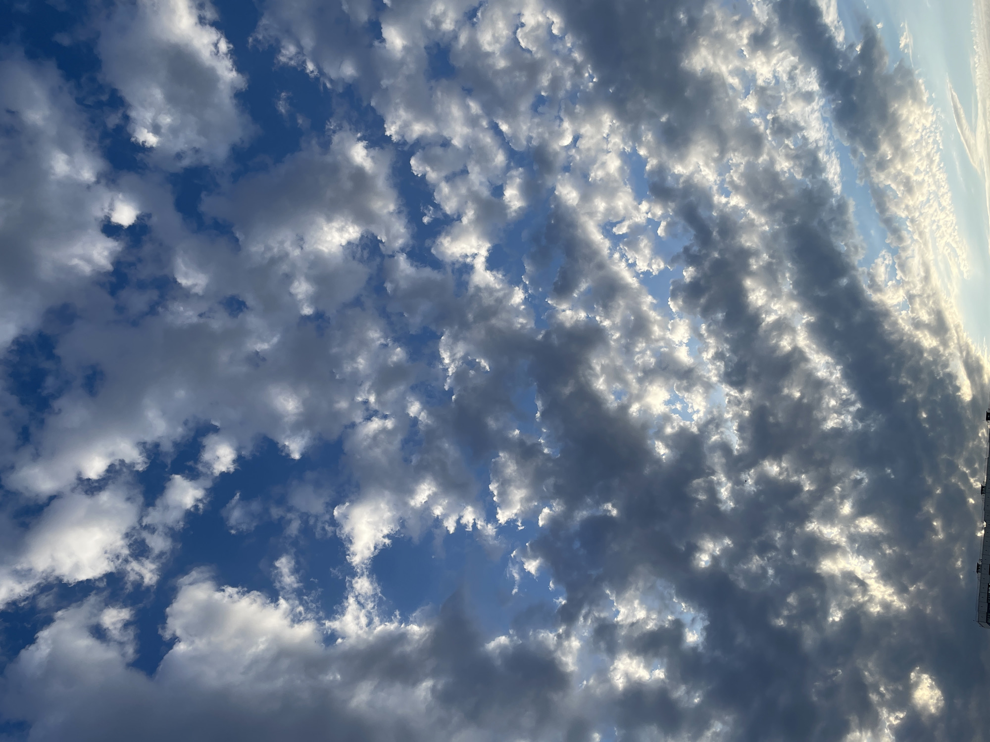 The image is a close-up, low-angle shot of a dramatic sky filled with a mix of dark gray and bright white clouds against a vibrant blue sky. The clouds are textured and appear to be moving, with sunlight filtering through them in places, creating a luminous effect. At the very bottom of the frame, the top of a modern, white building with multiple floors and visible windows can be seen, suggesting an urban environment. The building's architecture is simple and functional. The overall impression is of a dynamic and somewhat moody sky over a city. The location is identified as Rostov-on-Don, Russia.
