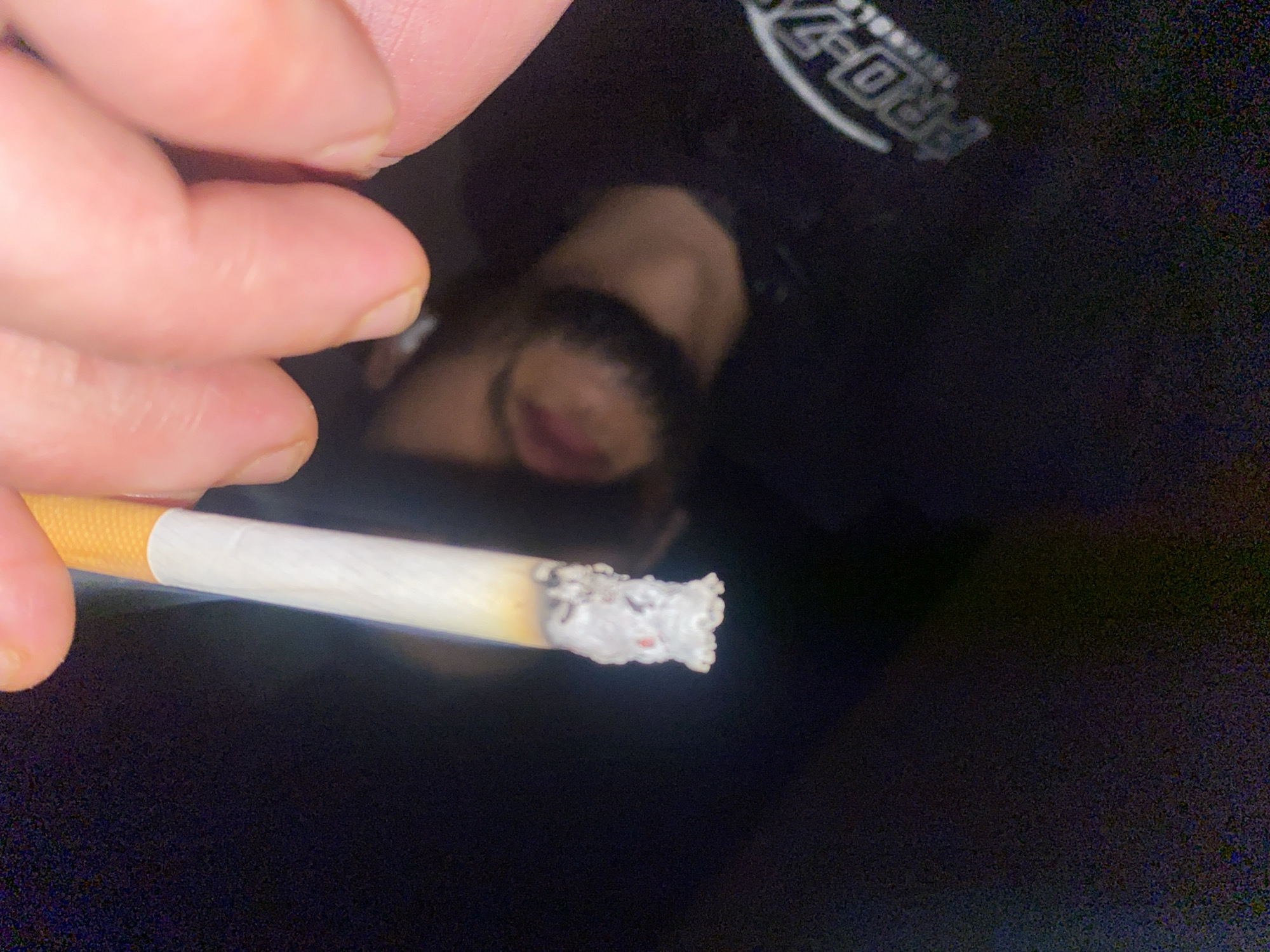 The image, taken in Schiltigheim, France, presents a close-up view of a human hand holding a lit cigarette. The cigarette features a tan filter and a white paper wrap, with a substantial column of white ash accumulated at its tip, displaying visible glowing embers and reddish-orange points of incandescence. The fingers of the hand, showing a light skin tone and visible fingernails, grip the cigarette near the filter. The background consists predominantly of a dark, reflective surface. This surface shows a blurry reflection of a human face, characterized by reddish lips and a dark beard or stubble, positioned centrally in the lower half of the frame. Additionally, a partial, curved reflection of a logo, displaying "PRO-Z" in white text on a dark background, is visible in the lower right portion of the reflective surface. The overall environment appears dimly lit.