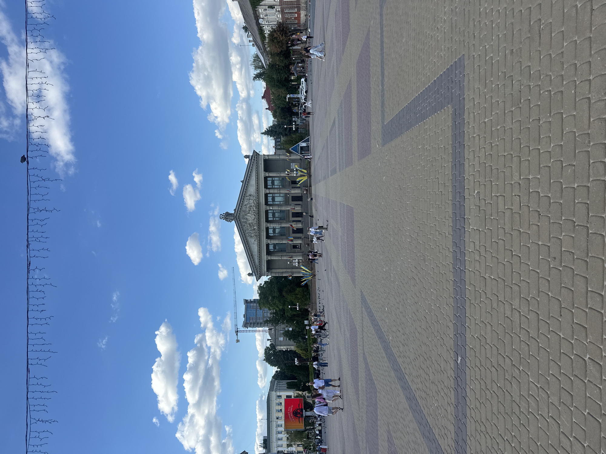 In Ternopil, Ukraine, a wide pedestrian plaza paved with interlocking bricks, featuring patterned sections of darker bricks, is the setting. A large, classical-style building with columns and an ornate facade dominates the background, displaying Ukrainian flags. To the left, a modern building with a large screen showing an image of a person and text is visible. Several people are dispersed across the plaza, some walking, some standing, and a group of pigeons is gathered on the ground. The sky is blue with scattered white clouds. Overhead, a string of decorative lights hangs across the scene. A construction crane is visible in the distance behind some buildings.