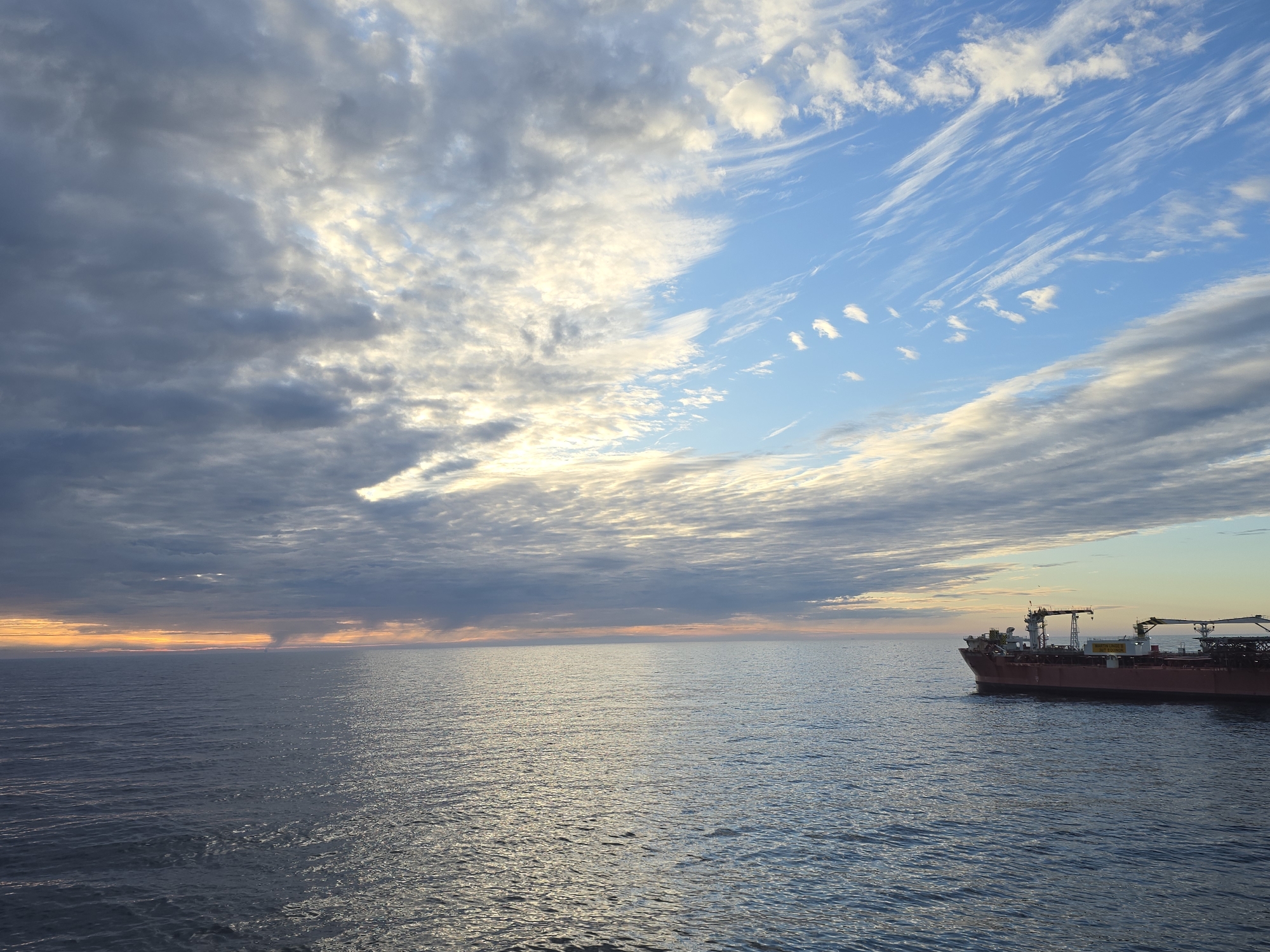 The image depicts a body of water, possibly an ocean, with a large ship on the right side. The sky is filled with clouds, transitioning from darker, gray clouds on the left to lighter, blue clouds on the right. The sun appears to be setting on the left horizon, casting a golden glow. The exact location of this scene is unknown.