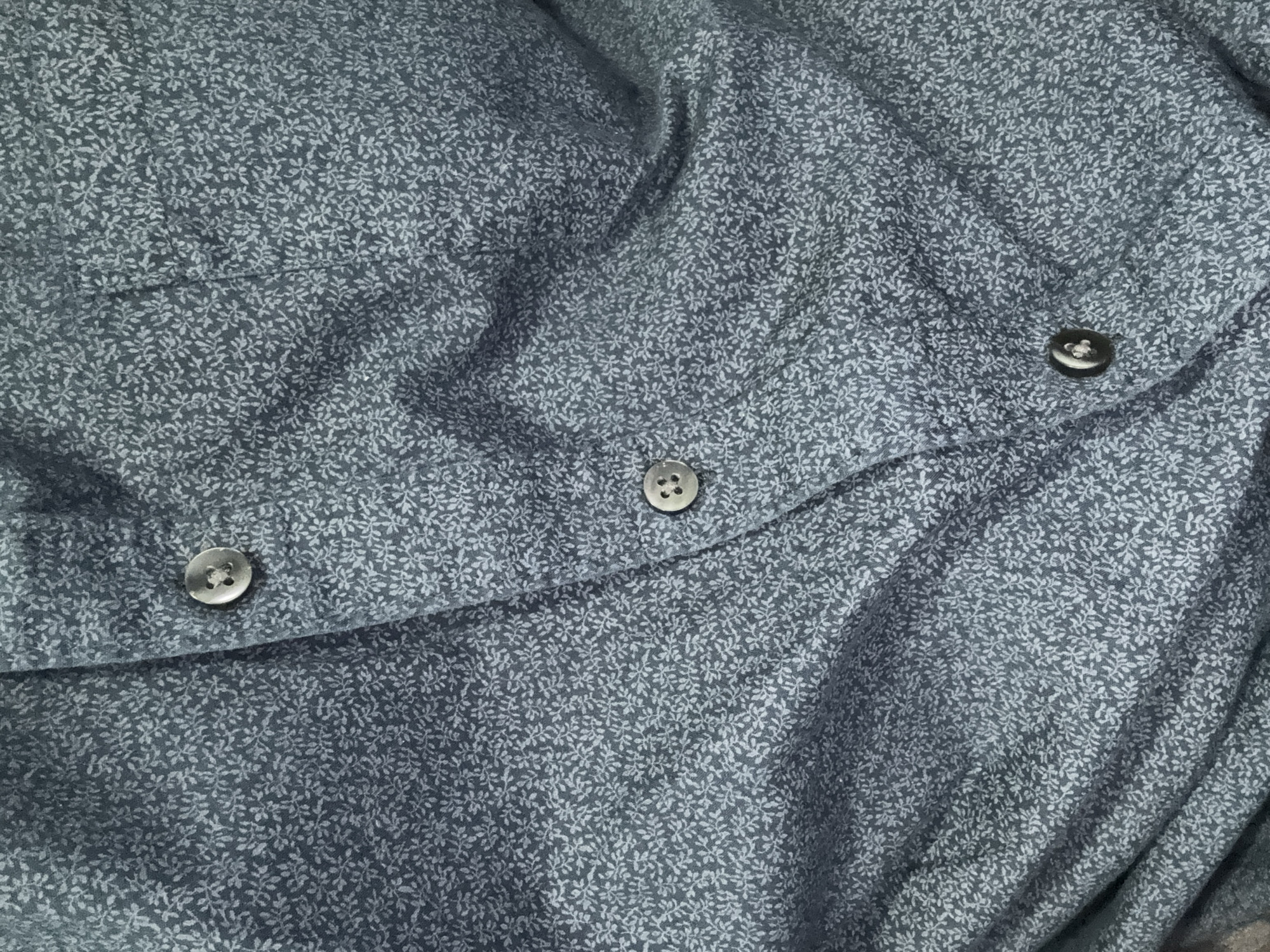 The image shows a close-up of a gray shirt with a small floral pattern. Three dark-colored buttons are visible, holding the fabric together. The material is slightly wrinkled. No location information for Cuautitlán Izcalli, Mexico, or any other location is discernible from the image. No people, subjects, actions, or interactions are visible.