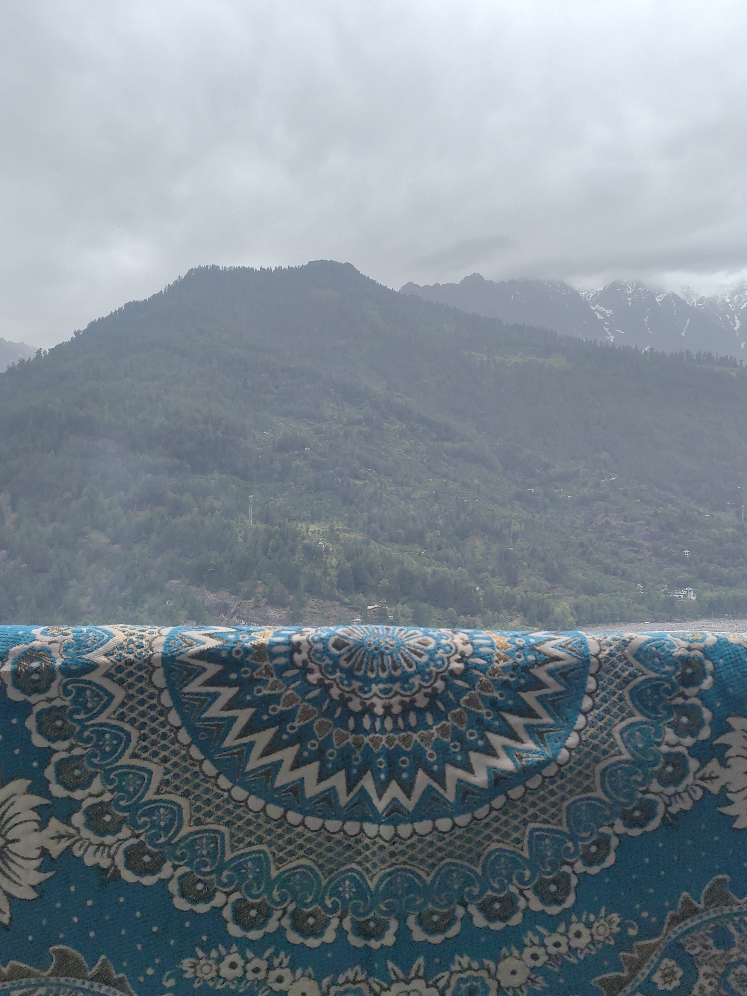 The image shows a view of mountains from what appears to be Vashisht, India. In the foreground, a blue textile with a white and brown intricate pattern is draped, partially obscuring the view. The textile has a symmetrical, mandala-like design with floral and geometric elements. Beyond the textile, a lush green mountain range rises, covered with dense trees. The peaks of some mountains in the distance are capped with snow, and the sky is overcast with clouds. The scene suggests a cool, possibly humid, day in a mountainous region.