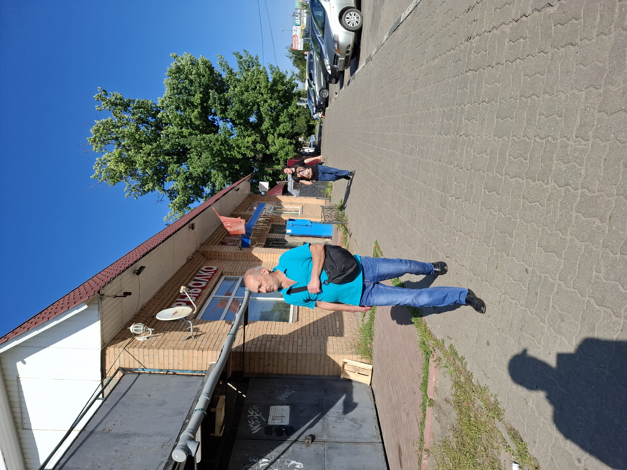 A man in a turquoise polo shirt and blue jeans walks down a paved sidewalk in front of a brick building. He carries a black satchel across his body. Behind him, another person wearing a black t-shirt and blue jeans walks away from the camera. The building has large metal doors and a sign with red lettering. A large green tree stands to the right of the building, and several cars are parked along the street. The sky is clear and blue.