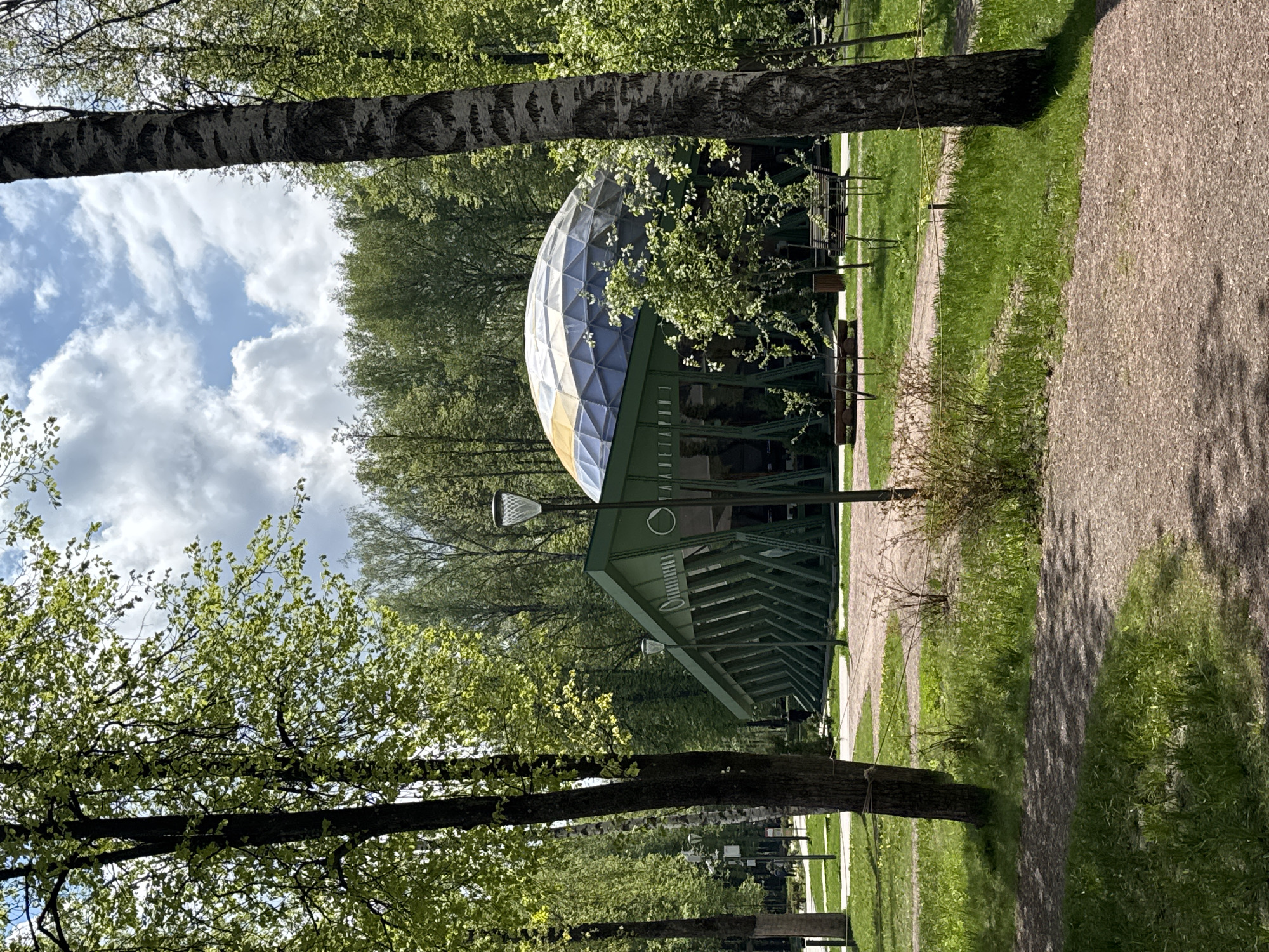 The image depicts a geodesic dome structure in a park setting. The dome has a segmented pattern, with a mix of silver, yellow and blue panels. It is supported by a green, angular framework that extends to the ground. Trees with fresh green leaves frame the structure, and a gravel path runs alongside it. The scene is well-lit, suggesting a sunny day. There's a clear sky with some clouds visible above the trees behind the dome. The location is the Nizhny Novgorod Planetarium, Russia.