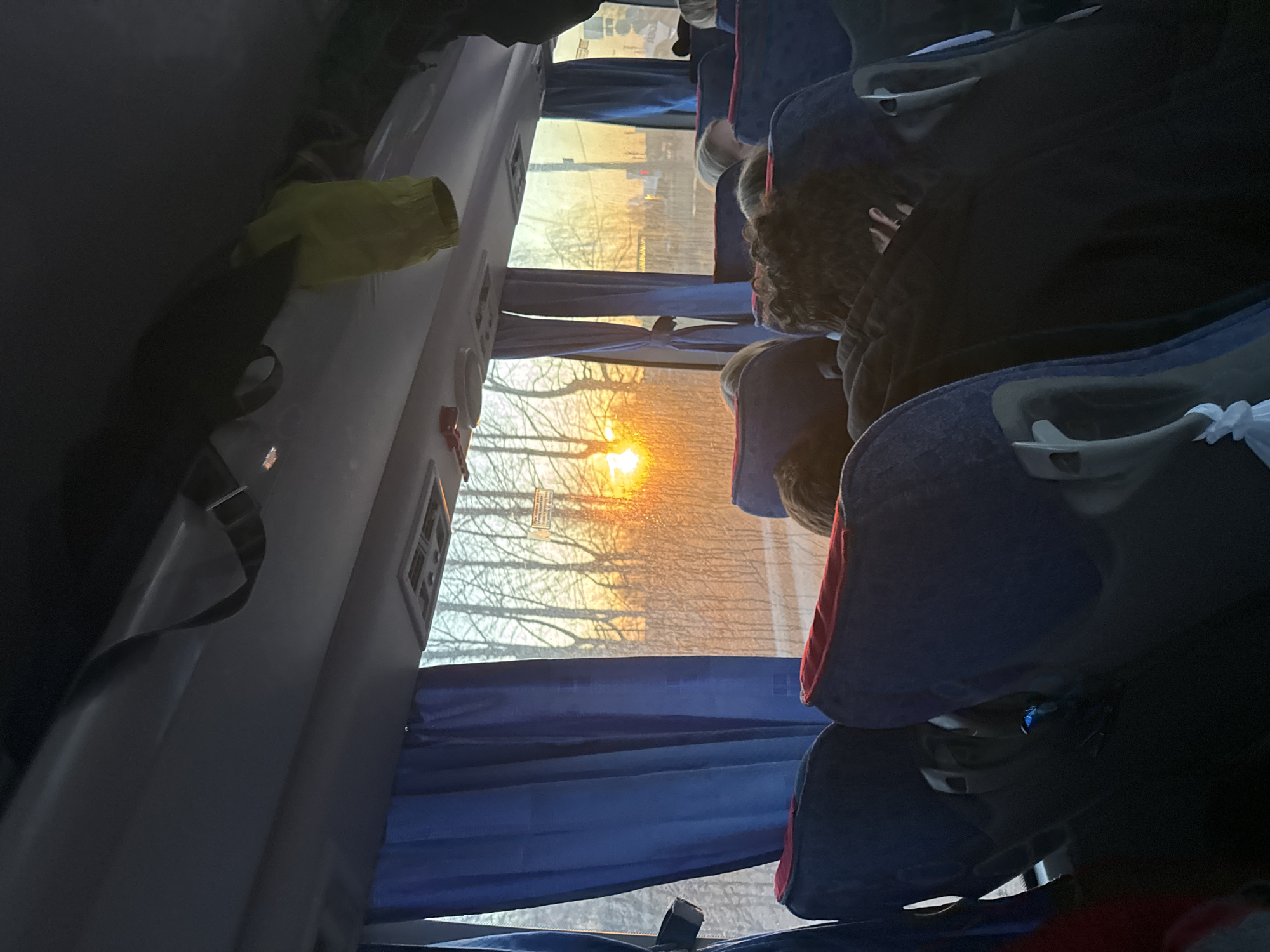 The image depicts the interior of a bus at sunrise. Sunlight streams through the window, silhouetting trees. Several rows of seats are visible, some occupied by passengers. Blue curtains partially cover the window. A yellow garment and a black strap hang on the side of the bus. It is not possible to determine the location as Osa, Russia from the provided image.
