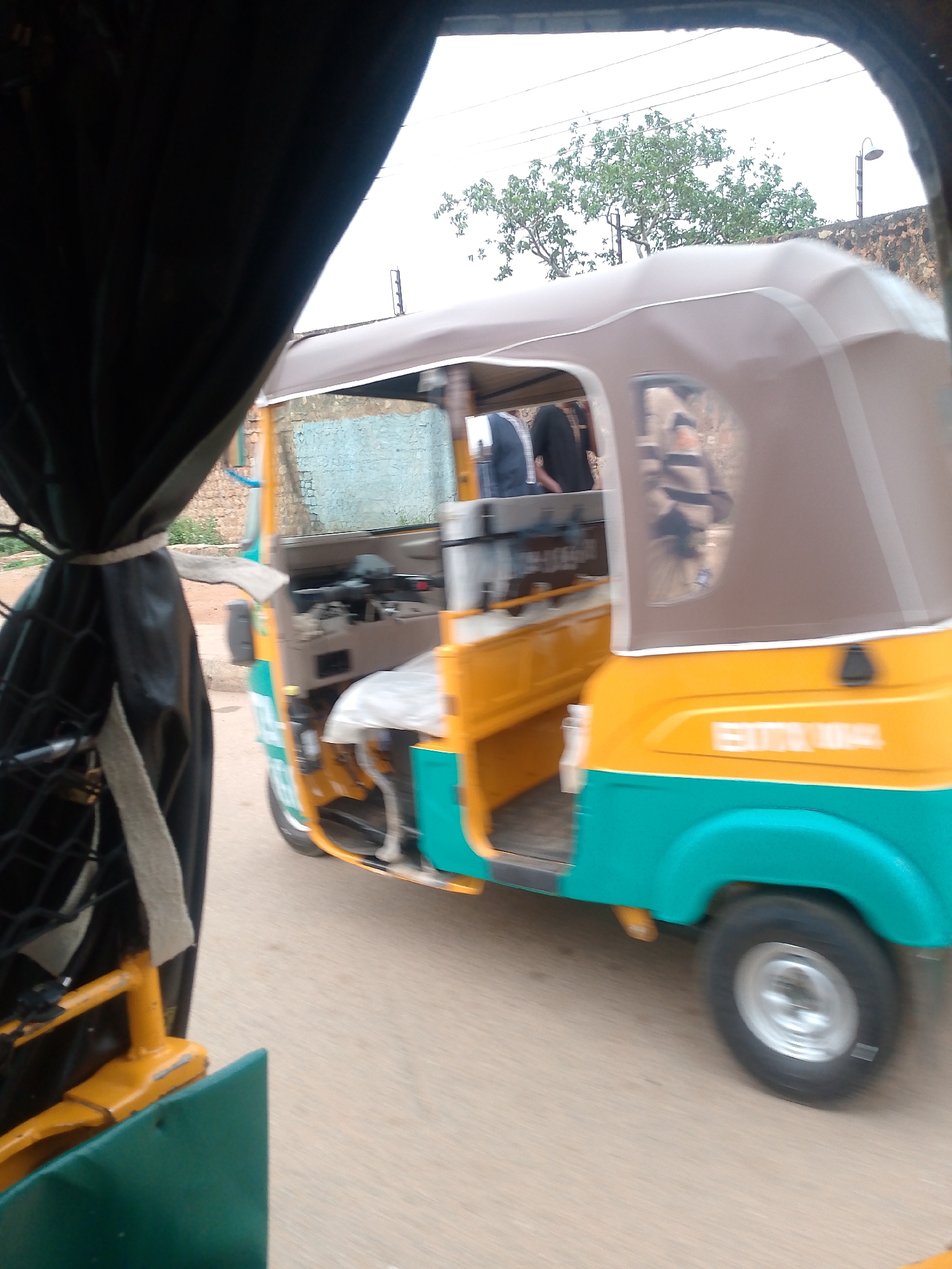 The image depicts a three-wheeled motorized vehicle, commonly known as a "keke," moving on a road, likely in transit in Bukuru, Nigeria. The keke in view is predominantly yellow with a green lower body and a brown canopy. There are passengers visible inside the keke. The camera perspective appears to be from inside another similar vehicle or structure. In the background, trees and buildings are visible.