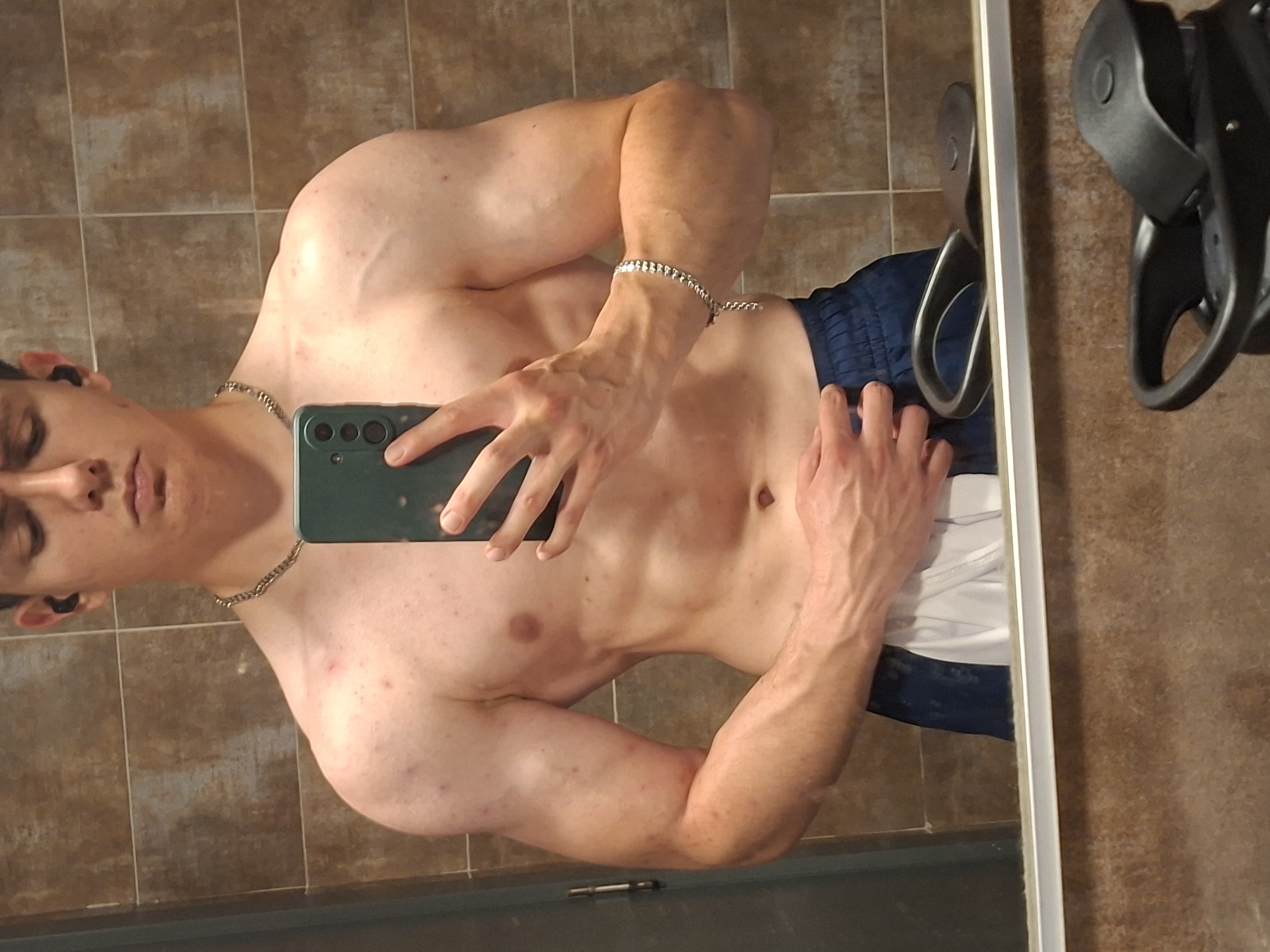 A shirtless young man with light skin is taking a selfie in front of a mirror in a tiled room, possibly a bathroom. He is wearing a silver chain necklace, a silver bracelet on his left wrist, and dark-colored earbuds. He is holding a dark green cell phone with his left hand. The man is also wearing blue and white shorts. A black object, possibly workout equipment, is visible in the upper right corner of the image. The location is not identifiable as Plovdiv, Bulgaria based on the image.