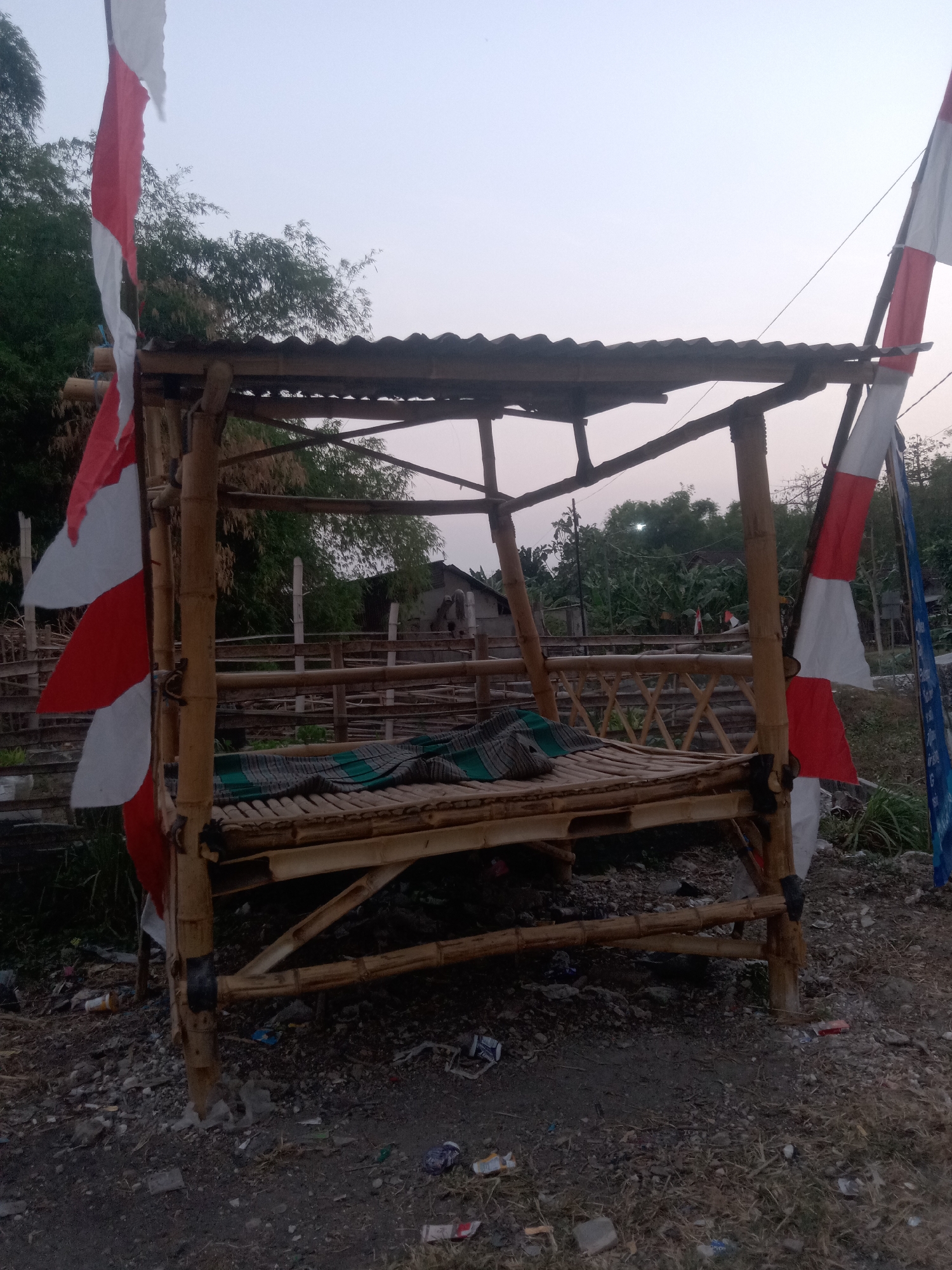 A medium-shot, eye-level, outdoor, full-color, slightly hazy photograph shows a simple, open-air bamboo structure with a corrugated metal roof, likely a gazebo or a resting place. Two Indonesian flags, red and white, are attached to poles on either side of the structure. The flags are tattered and appear to be weathered. The bamboo structure itself is rustic, with visible bamboo poles and woven bamboo panels for the railing. A woven mat or blanket, in shades of green and gray, is laid out on the bamboo platform, suggesting it is intended for seating or resting.
The ground around the structure is covered in loose gravel, dirt, and debris, including various pieces of litter such as plastic bottles and wrappers, indicating a somewhat neglected environment. In the background, a wooden fence is visible, and beyond that, a dense collection of trees and foliage, suggesting a rural or semi-rural setting. A few simple buildings can be seen in the distance among the trees, partially obscured. The sky is a pale, hazy gray, consistent with either dawn or dusk. There are no people visible in the image. The overall impression is one of a simple, utilitarian structure in a somewhat untidy, natural environment. The location is identified as Jumput, Indonesia, though this specific context is not visually evident in the photograph itself, which presents a generic rural Indonesian scene.