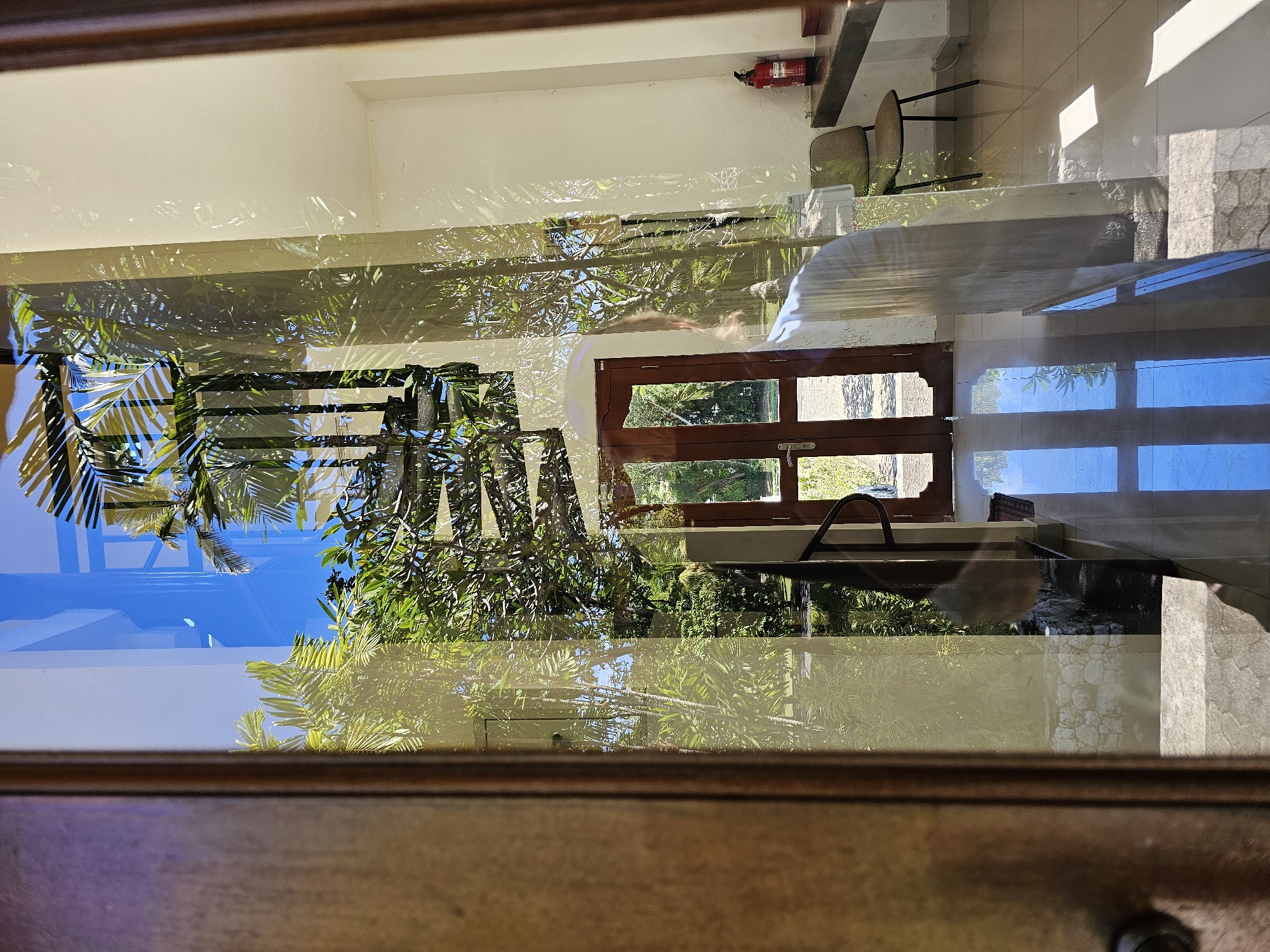 The image appears to be taken from inside a building, looking out through a window. Reflections on the window make it difficult to discern the scene clearly. The exterior features green foliage and blue sky, suggesting an outdoor setting with vegetation. Inside the building, the image captures a room with white walls, a fire extinguisher mounted on the wall, and furniture including chairs and possibly a table. A partially visible brown wooden door suggests a pathway to another area. The presence of reflections indicates a glass or reflective surface between the camera and the exterior scene. It's impossible to confirm the exact location in Kuta Selatan without additional information.