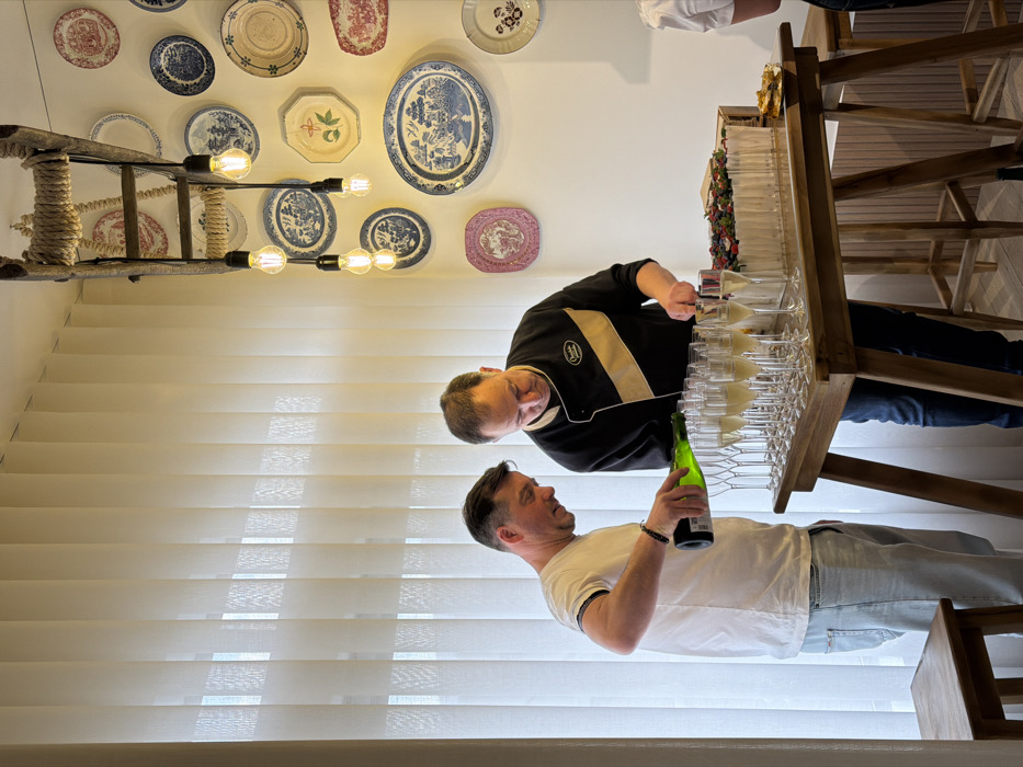 In Felgueiras, Portugal, two adult males are engaged in food and beverage preparation within an indoor commercial or event space. A male positioned on the left, wearing a white t-shirt and light blue jeans, is actively pouring a light-colored sparkling liquid from a green bottle into a row of multiple champagne flutes on a wooden high-top table. He has a dark bracelet on his left wrist. Immediately to his right, a male wearing a black chef's jacket with a tan diagonal stripe is cutting a large, rectangular cake with a silver knife. The cake is decorated with a top layer of mixed berries and has white, vertical, cylindrical elements along its sides. A collection of approximately 20 champagne flutes, many partially filled, are arranged on the table. Overhead, a decorative lighting fixture composed of a wooden ladder wrapped with rope suspends five bare Edison-style light bulbs. The background features a large window covered by vertical cream-colored blinds on the left and a white wall adorned with numerous decorative ceramic plates of various sizes and blue, pink, and white patterns on the right. A portion of a third individual's right shoulder and arm, wearing a white garment, is visible on the far right of the frame. A light-colored wooden stool is partially visible in the lower left foreground.