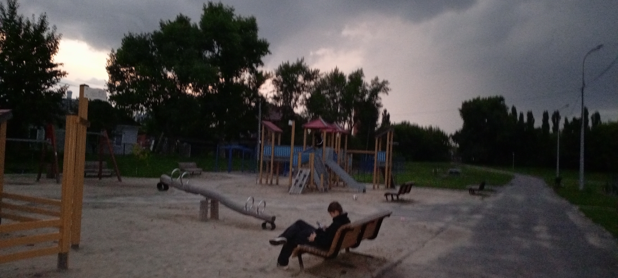 The image depicts a playground in Kirsanov, Russia, during dusk. The sky is overcast with dramatic clouds. In the foreground, a person sits on a park bench, looking down at a mobile device. The playground features a seesaw, a slide, and other play structures, constructed from wood and metal. A paved path runs through the scene, bordered by grass and trees. The overall atmosphere is somber, enhanced by the dim lighting and the approaching twilight.