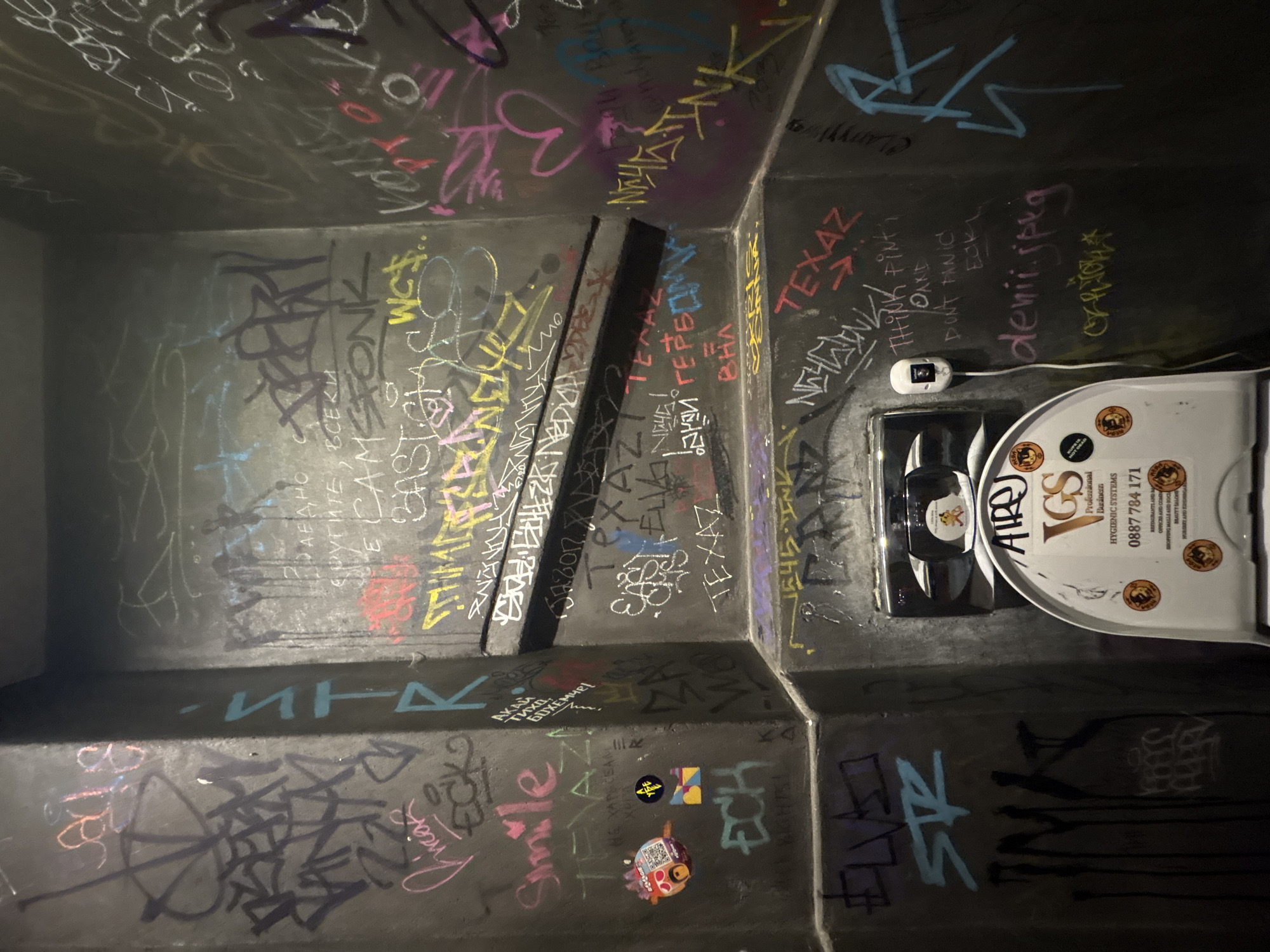 This image captures a public restroom in Sofia, Bulgaria, characterized by dark, graffiti-covered walls. The walls appear to be constructed from concrete or a similar material and form an angular, non-planar enclosure.
The walls are extensively covered with spray paint and chalk graffiti in multiple colors, including black, white, pink, yellow, red, and light blue. Various tags and stylized lettering are present, some in Cyrillic script (e.g., "АКАЙ ТИХА БОХЕМА!", "ДАЕХНО", "ВСЕКИ", "ТЕРАЗ") and others in Latin script (e.g., "Smile," "TEXAZ," "ECH," "denii.jpeg," "THINK PINT AND DON'T PANIC"). A pink heart is visible on the upper right wall section. Two small stickers are affixed to the upper left wall: a colorful circular QR code sticker labeled "Space" and a smaller square sticker with geometric patterns and "YEE" text. Two dark, horizontal ledges or shelves divide the upper and middle sections of the back wall.
In the lower center of the image, a white, wall-mounted toilet seat cover dispenser with a metallic top is visible. A prominent "VGS Professional Business Hygienic Systems" sticker is affixed to its front, displaying a phone number (0887 784 171) and listing various types of facilities served. Several smaller circular stickers featuring a cartoon animal and text like "CLIPS IS MY OES" are also on the dispenser. Reflected in the metallic top of the dispenser is a small sticker of a cartoon teddy bear. To the right of the dispenser, on the wall, a white automatic hand sanitizer unit is mounted, connected by a visible white cable.
The lighting is dim and artificial, creating shadows and highlighting the texture and graffiti of the walls. No individuals are present in the scene, and no actions are depicted. The overall environment suggests a restroom within a bar, club, or similar entertainment venue in Sofia.