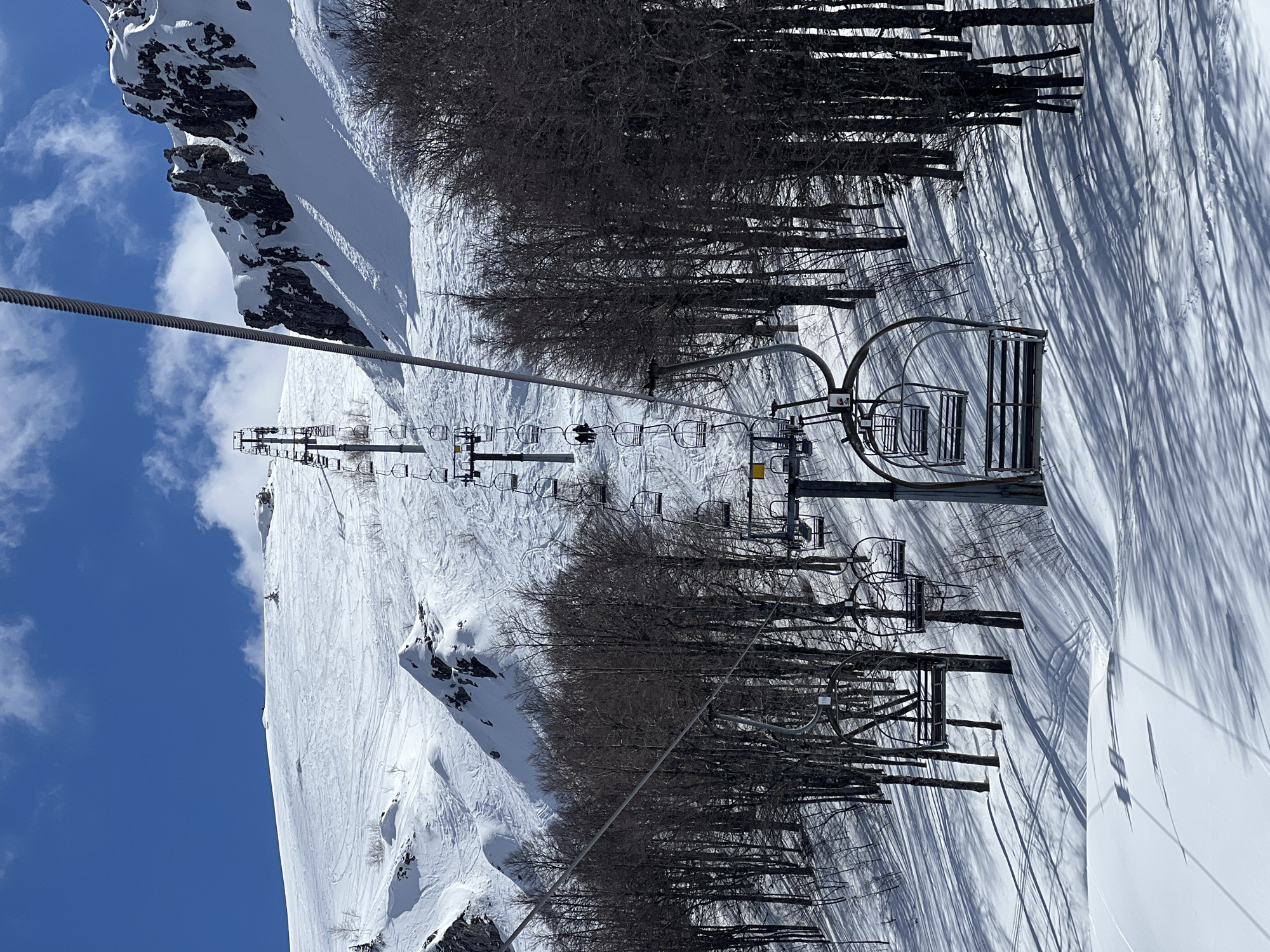 The image depicts a snowy mountain landscape with a ski lift in operation. The location is Katun Bačko brdo, Montenegro. The sky is blue with scattered clouds. The ground is covered in snow, showing ski tracks. There are bare trees alongside the ski slope, casting long shadows on the snow. The ski lift consists of chairs suspended from a cable. The ski lift structure and chairs are visible. The image is taken from a high vantage point, looking down the mountain.