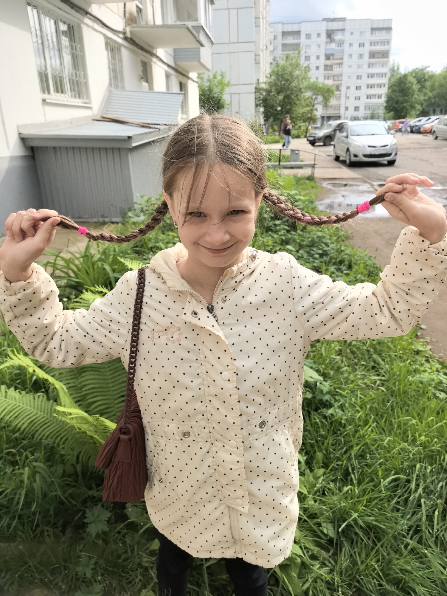 A young girl stands outdoors, holding the ends of her braided pigtails up in her hands. The braids are secured with pink hair ties. She has a slight smile on her face. She is wearing a white polka-dot jacket, black pants, and a brown purse with a braided strap. She is standing in an area with green grass and ferns. Behind her is a residential area with apartment buildings, trees, and several parked cars. A person walks along a sidewalk in the background. The image was likely taken in the city of Chaikovsky, Russia.