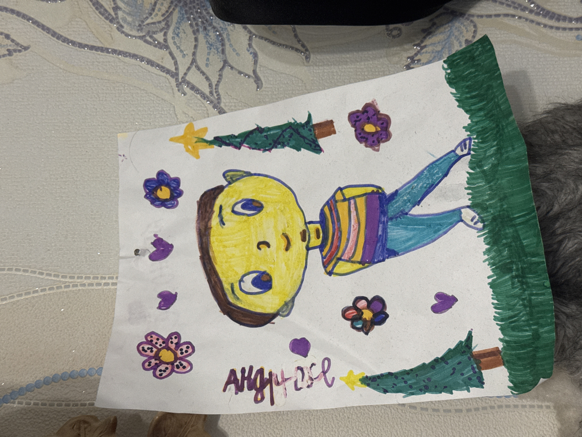 The image shows a child's drawing on white paper. The drawing includes a yellow-faced figure with brown hair, wearing a striped shirt, and blue pants. There are flowers and heart shapes drawn around the figure. At the top and bottom edges are stylized green fir trees. The drawing appears to be signed with the name "AHGPOSE". The background is a light-colored patterned surface with some indistinct objects and a section of dark gray fur or fabric. The image was likely taken in Chuprino, Russia.