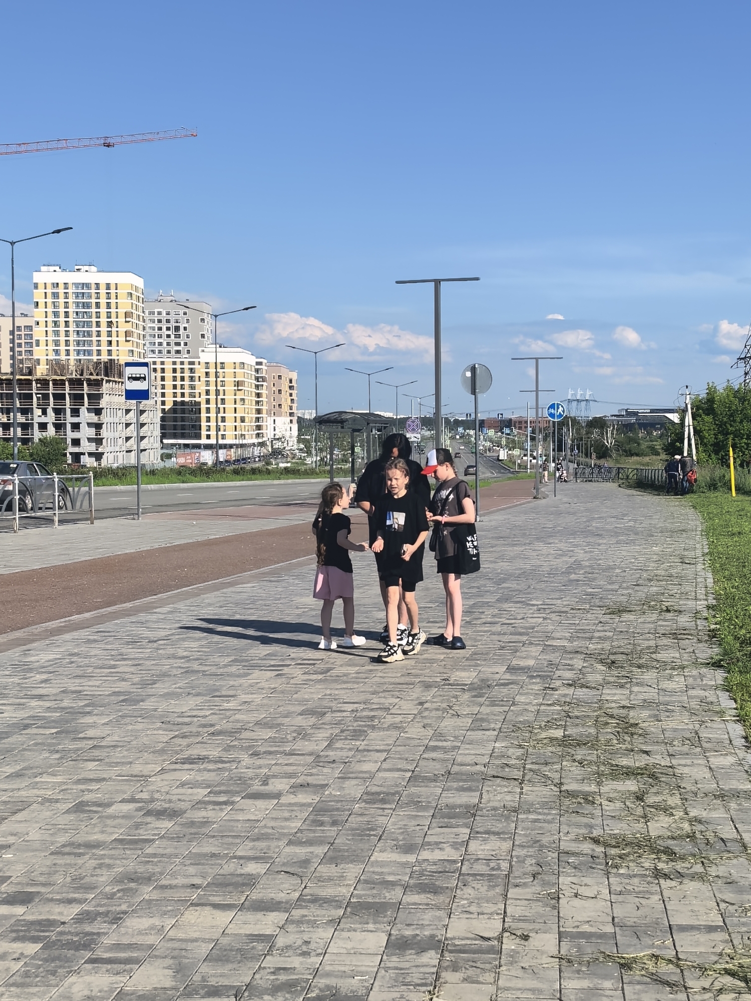 The image shows a group of four people, three young girls and one adult, standing on a paved walkway. The girls are in the foreground, with the adult slightly behind them. The girl on the left wears a black t-shirt and pink shorts. The girl in the middle wears a black t-shirt and black shorts, and has a bag with some text on it. The girl on the right wears a black t-shirt and dark shorts. The adult, partially obscured, is wearing a black t-shirt and black shorts. They appear to be walking or standing around, possibly waiting.
The setting is an urban environment with modern buildings in the background, some of which appear to be under construction. There is a street with a few cars visible, and a bus stop sign is also present. Overhead, a construction crane is visible against a clear blue sky with a few scattered clouds. The weather appears to be sunny and warm, suggesting daytime. The overall atmosphere seems casual and relaxed. The pavement is made of large, grey rectangular tiles. To the right of the walkway, there is a grassy area. The image is taken from a slightly elevated perspective, looking down at the subjects and the scene.