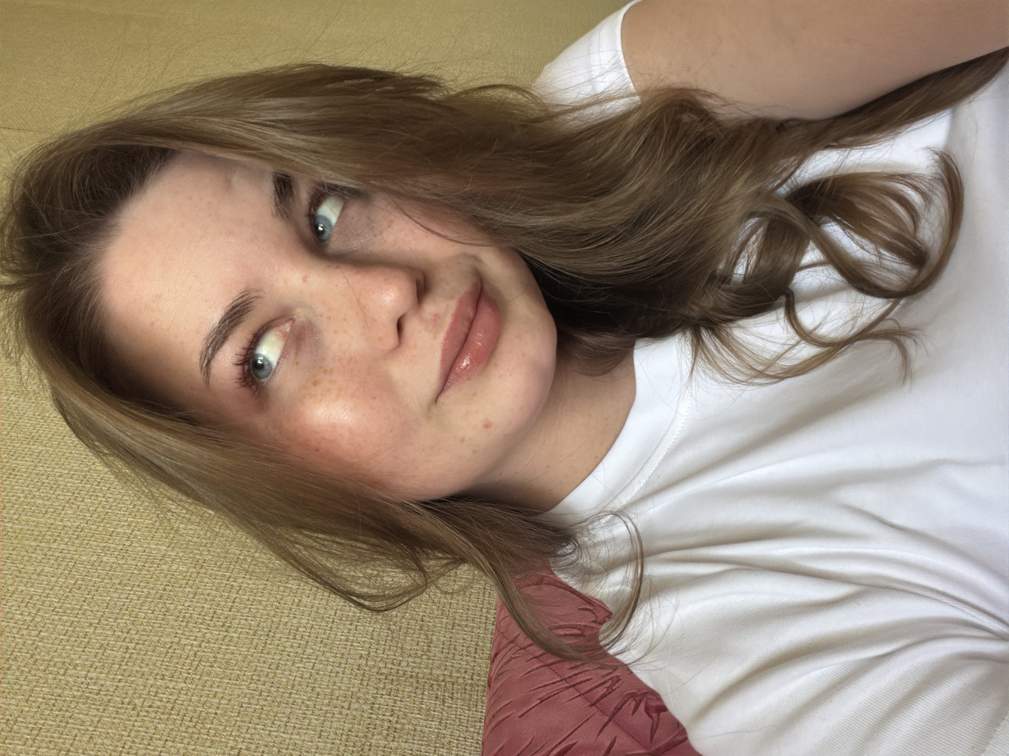 A young woman with fair skin and freckles is pictured. She has light brown hair with waves and blue eyes. She is wearing a white t-shirt. The background consists of a textured tan surface and a red fabric. The location in the city of Chaikovsky, Russia, cannot be determined from the image.