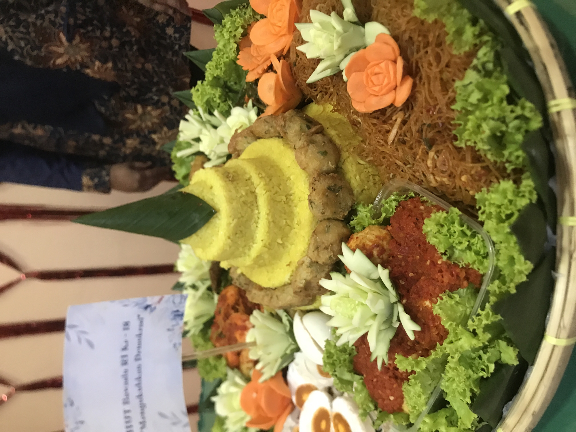 The image depicts a "tumpeng," a traditional Indonesian cone-shaped rice dish, elaborately decorated with various side dishes and garnishes. The tumpeng, the central element, is composed of yellow rice stacked in tiers, resembling a small mountain. Around the base of the rice cone, there are fried patties and other unidentified dishes, interspersed with carved vegetable flowers, most likely made from cucumbers or similar vegetables.
Additional decorations include orange-colored carrot roses, green lettuce, and what appear to be slices of salted duck eggs. Some food items are contained in a small, clear plastic container. The arrangement sits on a woven tray made of bamboo or similar material.
A small sign, likely commemorating an event, is visible to the side, and part of a person's clothing, consisting of a dark blue and brown patterned top, is seen in the background.
Given the presence of "tumpeng," a significant element in Indonesian culture and festivities, the location is likely in Indonesia. Considering the instruction to include the city, the scene might potentially be associated with an event or celebration in Lhokseumawe, Indonesia.