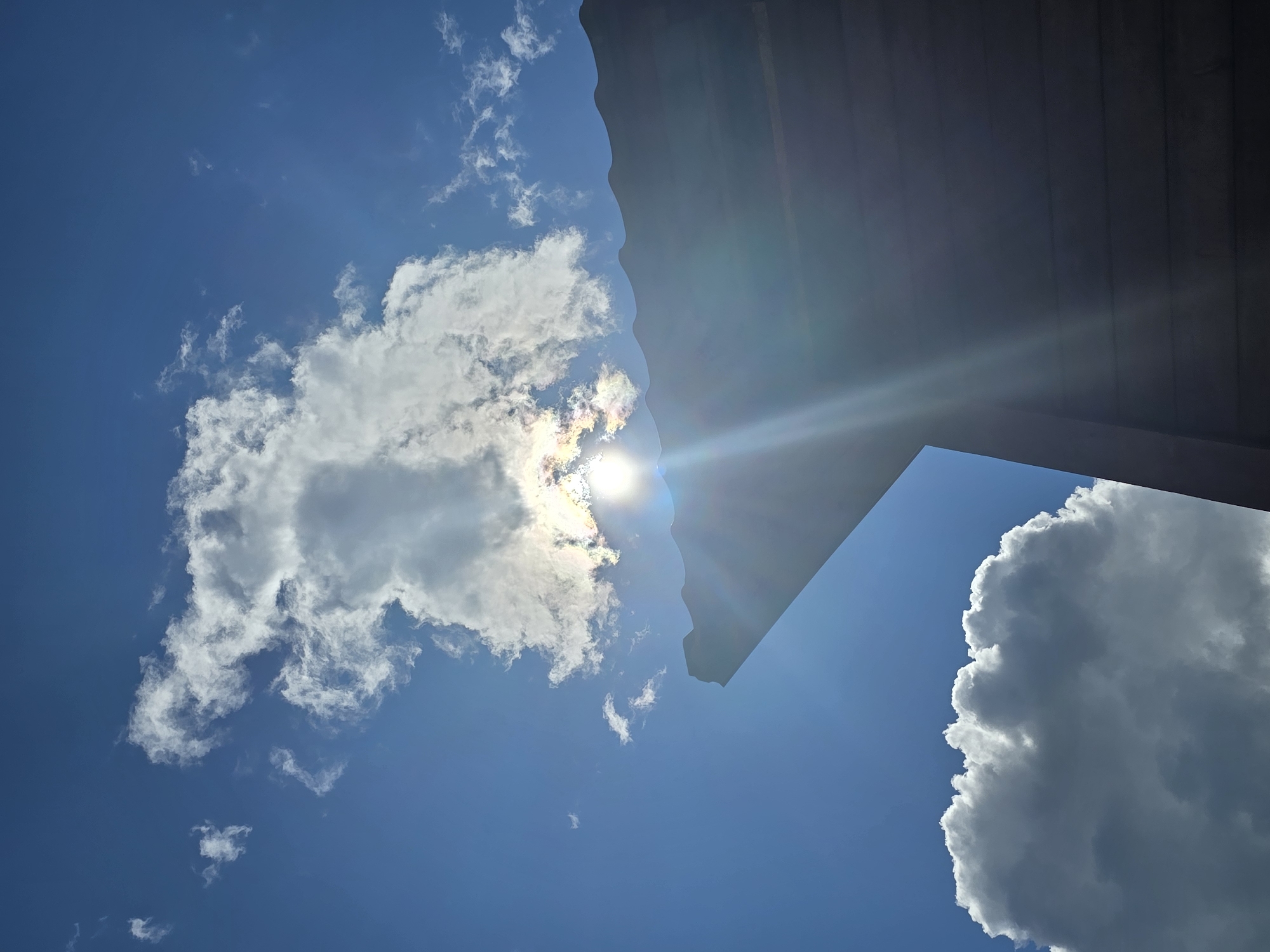 The image shows a bright sun partially obscured by a cloud, casting rays of light across the frame. The sun's glare is intense, creating a lens flare effect. In the foreground, the silhouette of a building's roofline is visible, with its dark, wooden siding horizontally oriented. The roof has a corrugated texture and a distinctive, angular edge. The sky is a vibrant blue, with several fluffy white clouds scattered across it. One large cumulus cloud is positioned in the lower left corner of the frame. The scene appears to be outdoors, likely in a residential or rural setting, given the presence of the building's roof. There are no people visible in the image. The overall impression is of a sunny day with dramatic lighting.