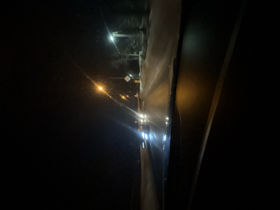 The image depicts a nighttime scene observed from the interior of a vehicle, looking forward through the windshield. The top portion of the dashboard is visible in the lower foreground. The environment is dark, suggesting night, with snow visible along the roadside.
A two-lane paved road stretches into the distance, illuminated by multiple streetlights and oncoming vehicle headlights. Several vehicles are approaching from the opposite direction, their headlights appearing as streaked bright white and blue light sources due to motion blur or camera exposure.
On the right side of the road, snowbanks line the edge. A prominent streetlight with an orange glow is visible, along with a secondary streetlight emitting a cooler, whiter light further down. Adjacent to the orange streetlight, a yellow diamond-shaped road sign, indicating a priority road, is mounted on a pole. Below this sign, a reddish-orange object or structure is present. Trees, partially obscured by darkness and snow, are visible behind the roadside elements. No individuals are explicitly visible. The scene is located in the city of Maksimkovo, Russia.