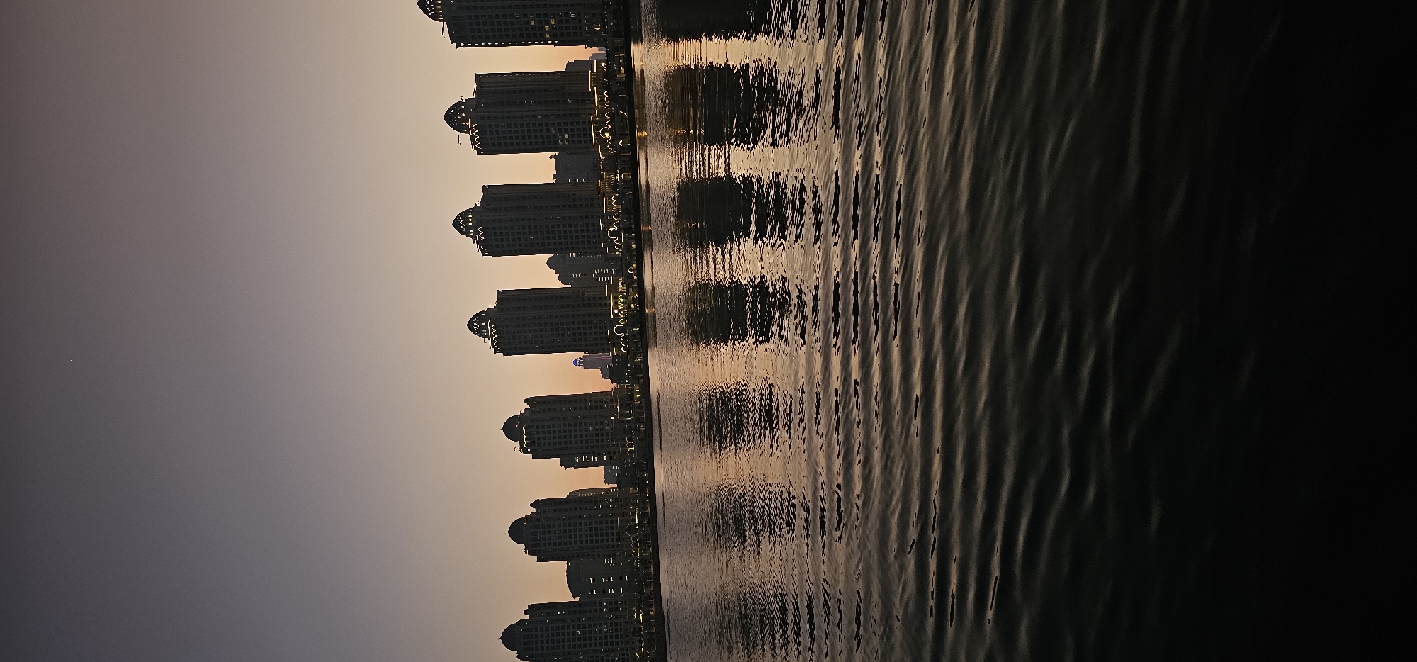 This media file depicts a waterfront cityscape at sunset in Doha, Qatar. The primary subjects are a row of identical, multi-story buildings with distinctive domed tops, lining the edge of a body of water. These buildings are silhouetted against a gradient sky that transitions from a pale orange at the horizon to a dusky purple and gray overhead. The water's surface is rippled, reflecting the warm glow of the sunset and the illuminated windows of the buildings, creating a shimmering, abstract pattern.
The scene suggests a serene evening ambiance. The time of day is clearly sunset, indicated by the warm, diffused light and the shift in sky color. The weather appears calm, with the water exhibiting only gentle ripples. The architectural style of the buildings, particularly the domes, suggests a modern, possibly resort-style development. While no people are directly visible, the presence of illuminated windows implies habitation or business activity within the buildings. The overall impression is one of urban elegance and tranquility, where modern architecture meets the natural beauty of the waterfront at dusk. No visible text is present in the image.