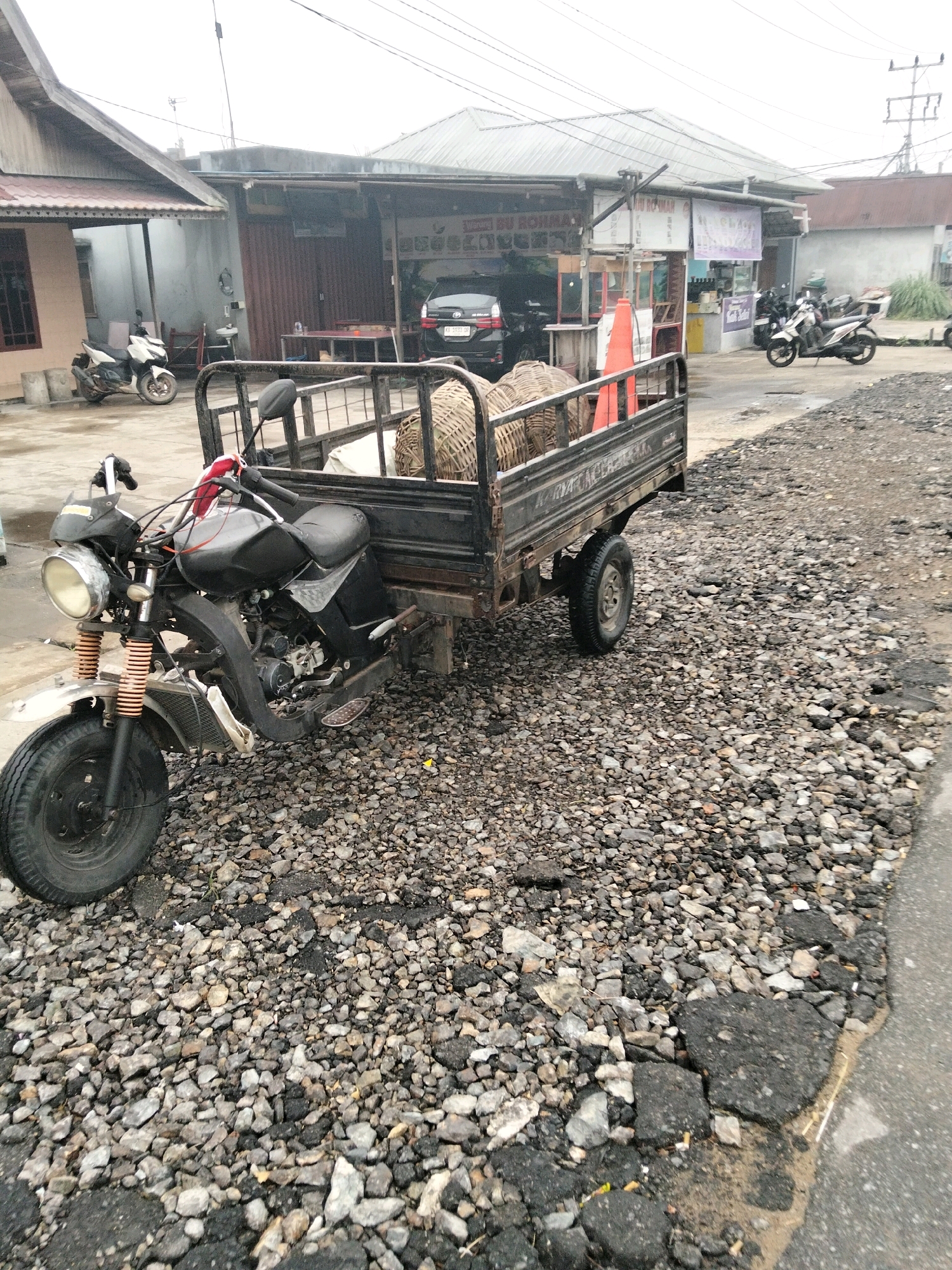 The image shows a three-wheeled motorcycle with a cargo trailer parked on a gravelly road in front of several buildings. The motorcycle is black and appears to be modified for hauling. The trailer is also black and contains several woven baskets. In the background, there are shops and houses, and a black car is visible. The sky is overcast, suggesting it may be a cloudy day. The scene is located in the city of Arang Limbung, Indonesia, as indicated by the license plate on the black car, which reads "KB 5533 DP," a format common for vehicles registered in Kalimantan, Indonesia.