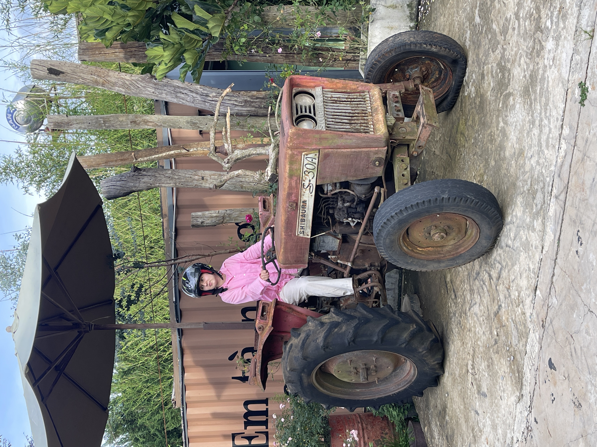 A person wearing a pink shirt, white pants, and a black helmet is sitting on a red, rusty tractor. The tractor is parked on a concrete surface. Behind the tractor is a container with the text "Em" visible on it. There is an umbrella providing shade over the tractor and the person. The background includes various plants, trees, and wooden poles. The photograph was taken in Thôn 5, Vietnam.