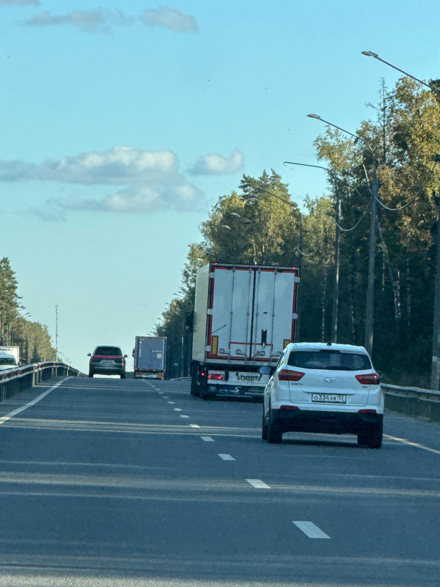An outdoor, eye-level, medium shot captures a bright, clear day on a highway in an unknown location in Russia. The road is paved with asphalt and marked with white lane lines. A white SUV is in the foreground, traveling in the right lane. Behind it, a white semi-truck with a blue trailer is in the left lane. Further ahead, a dark-colored SUV is in the left lane, followed by another blue trailer. Trees with green and yellow foliage line the right side of the highway, and light posts are visible along the road. The sky is a light blue with scattered white clouds.