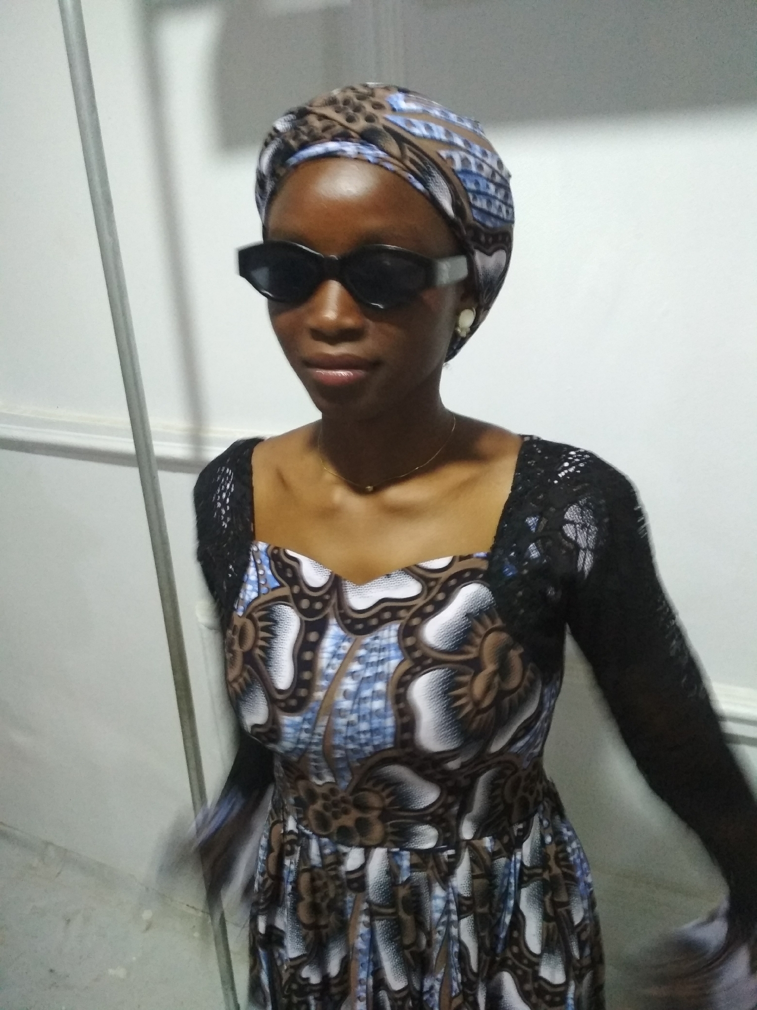 A woman stands indoors against a white wall, wearing a patterned headwrap, black sunglasses, and a dress with a mixed print bodice and black lace sleeves. The dress features a square neckline, and she is wearing a delicate gold necklace. A vertical metal bar is visible to the left of the frame.