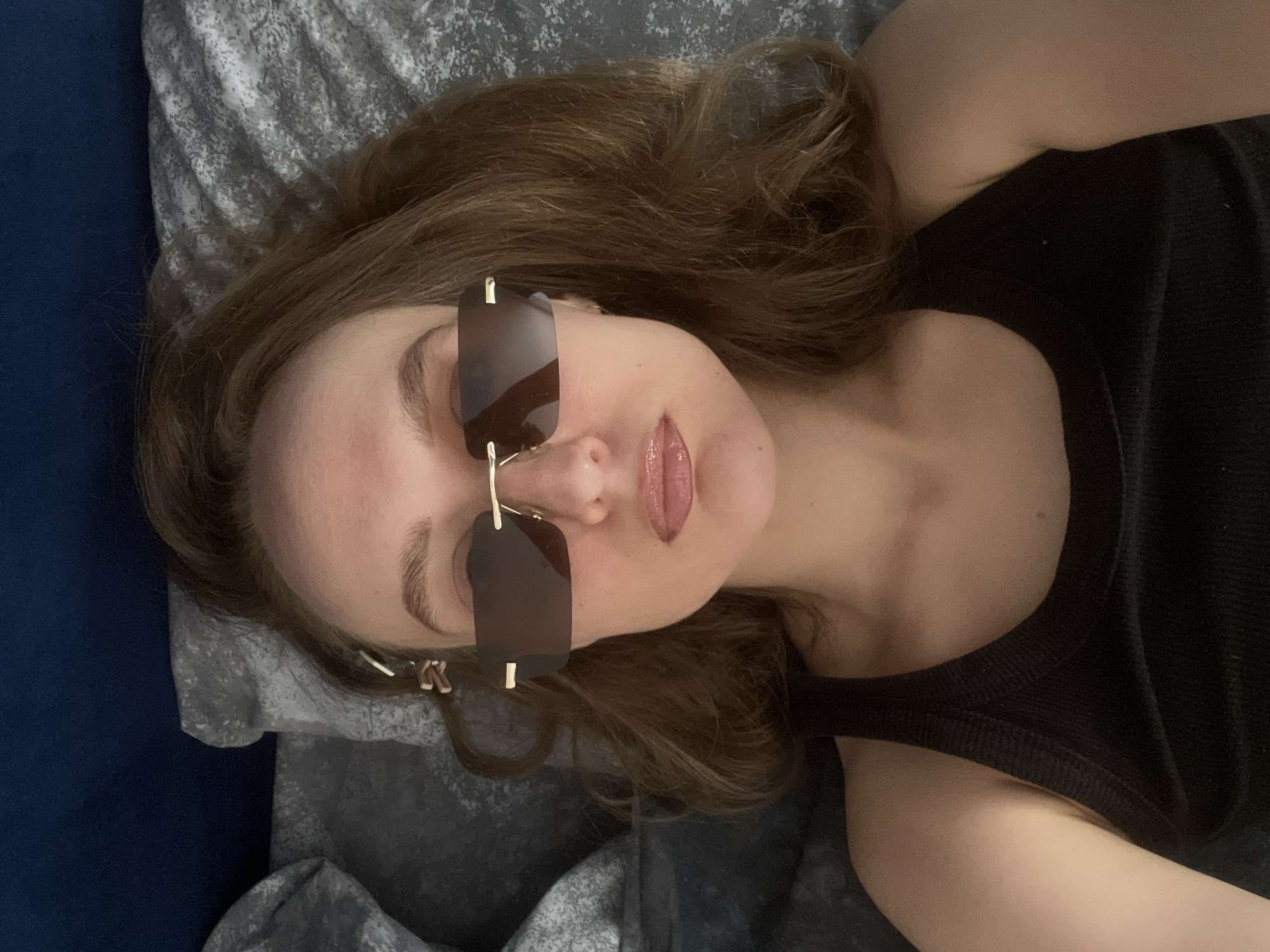 A woman with light skin and brown hair is lying on a bed with gray patterned bedding and a blue headboard. She is wearing a black tank top and rectangular sunglasses with brown lenses and gold frames. The location cannot be determined as the image does not show any geographical markers or landmarks.