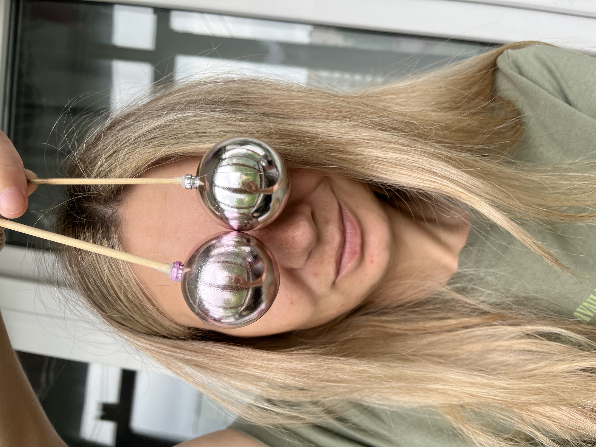 The image presents a close-up, rotated portrait of a person, likely a woman, with long, flowing blonde hair. The subject's head is tilted, and the image is oriented 90 degrees counter-clockwise.
**Subjects:** The primary subject is a woman with fair skin and long, straight, blonde hair, with some darker roots visible near the scalp. Her eyes are obscured by two large, spherical, highly reflective metallic ornaments, possibly decorative balls or Christmas ornaments, held on thin, light-colored sticks (resembling wooden skewers). Part of her face, including her nose and mouth, is visible below the spheres. She is wearing a green or olive-colored top.
**Activity:** The person is playfully posing, holding the two reflective spheres directly in front of her eyes, effectively covering them. This creates a humorous and whimsical effect.
**Scene/Setting:** The setting appears to be indoors, with a large window or glass door visible in the blurred background, allowing ample natural light. The spheres reflect distorted images of window frames and outdoor light, confirming the indoor-outdoor view. The context provided indicates the location is Chaikovsky, Russia.
**Notable Details:** The spheres are a prominent feature, appearing silvery with a subtle pinkish tint due to reflections. One of the sticks holding a sphere has a small purple decorative element near where it attaches to the sphere. The woman's lips are slightly parted, suggesting a relaxed or playful expression. The lighting is bright natural daylight. Indistinct, blurry text is partially visible on the green shirt, but not clearly legible.