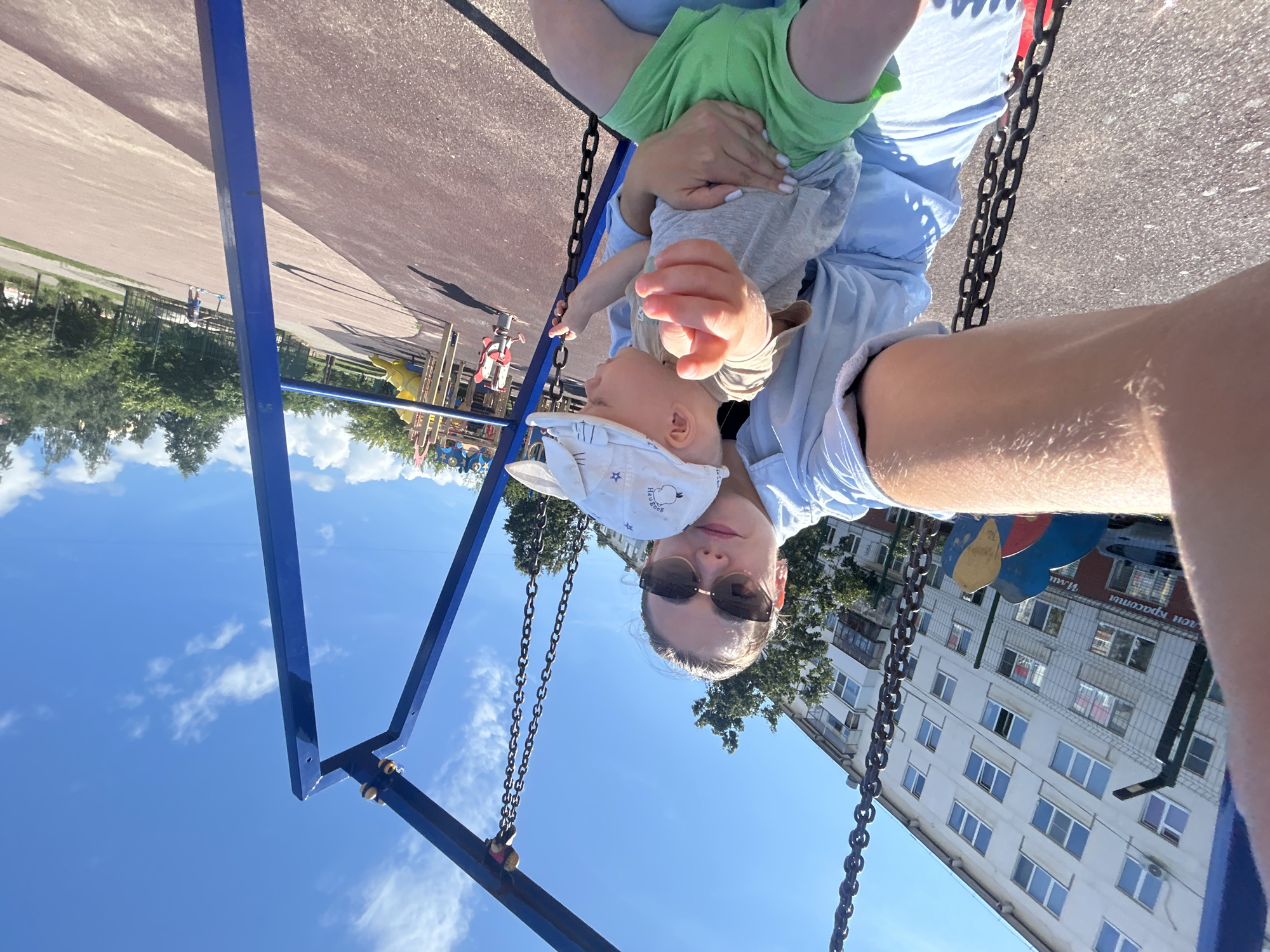 A woman is taking a selfie on a swing set at a playground. She is holding a young child on her lap. The child is wearing a white hat and green shorts, and a tan t-shirt. The woman is wearing sunglasses and a light blue shirt. In the background, there is a multi-story building, trees, and other playground equipment. The ground of the playground appears to be rubberized. The sky is blue with scattered white clouds. The location is the playground located on Lenina Street in Chaikovsky, Russia.