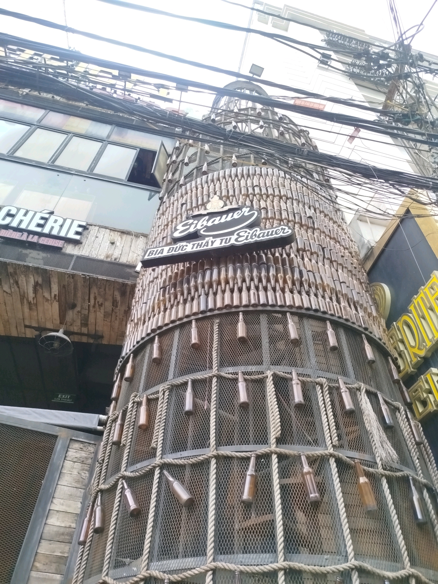 The image depicts a cylindrical structure covered in numerous brown glass bottles, which appears to be part of a building's exterior in an urban setting. Above the bottle-covered structure, the facade of a building with glass windows is visible, along with a sign that reads "CHERIE" and "BIERE A LA CERISE." A prominent sign on the bottle structure says "Eibauer" with Vietnamese text below it. The scene is characterized by an abundance of overhead electrical wires, suggesting a densely wired urban environment. The bottom portion of the structure features a metal mesh enclosure with ropes and more glass bottles attached. A sign reading "EXIT LOI THOAT" is visible in the lower left. The overall impression is of a commercial establishment with a unique, bottle-themed architectural element. This specific establishment is located on **Phan Dinh Phung Street in Da Lat, Vietnam**.