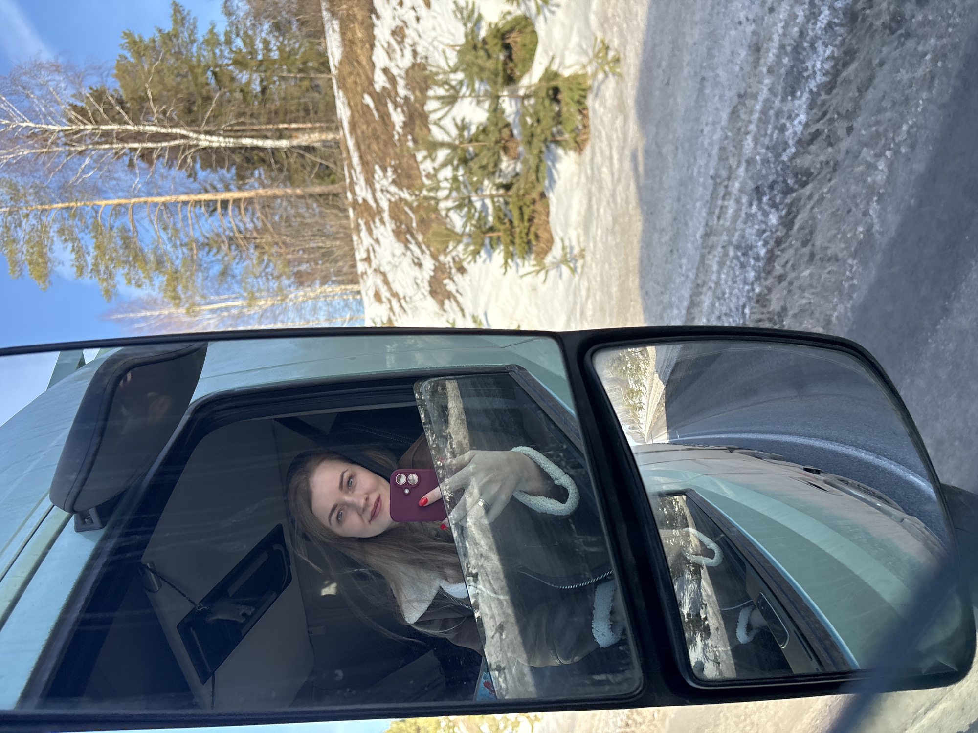This image depicts a scene in Bannoe, Russia, viewed from inside a light-green vehicle, likely a truck cab, with a significant portion of the composition focused on the driver's side window and mirror. A light-skinned female with long, light brown hair, wearing a brown jacket with white fleece lining, is visible through the open passenger window. She holds a dark red smartphone with a visible dual-lens camera system and is actively taking a photograph, her gaze directed towards the camera. Her left hand, adorned with red nail polish and a silver-toned ring on the ring finger, holds the phone.

The environment outside the vehicle is a natural, partially snow-covered landscape under a clear blue sky. A hillside with patchy snow cover and brown vegetation rises in the background. Scattered evergreen trees, appearing to be pines, stand alongside bare deciduous trees, possibly birches. Several small, green evergreen saplings are visible on the lower snowy slope. The vehicle's large side mirror reflects a segment of the truck's light-green body and a section of an asphalt road with visible white lane markings, flanked by icy or slushy snow. The overall impression is a winter or early spring setting.