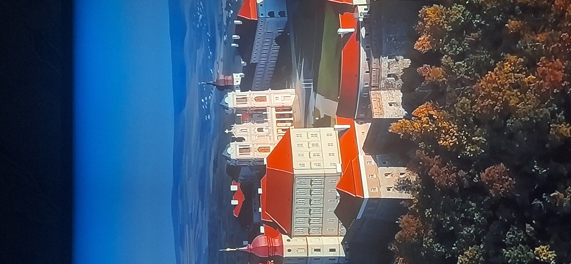 This is an aerial, wide-angle, vertical photograph of the Arboretum "Oleksandriya" in Bila Tserkva, Ukraine. The image captures a complex of buildings with red-tiled roofs, including what appears to be a palace or church with ornate white facades and twin towers. The buildings are surrounded by trees exhibiting autumnal foliage in shades of red, orange, and yellow. The background shows a hazy landscape with rolling hills and a clear blue sky. There are no people visible in the image.