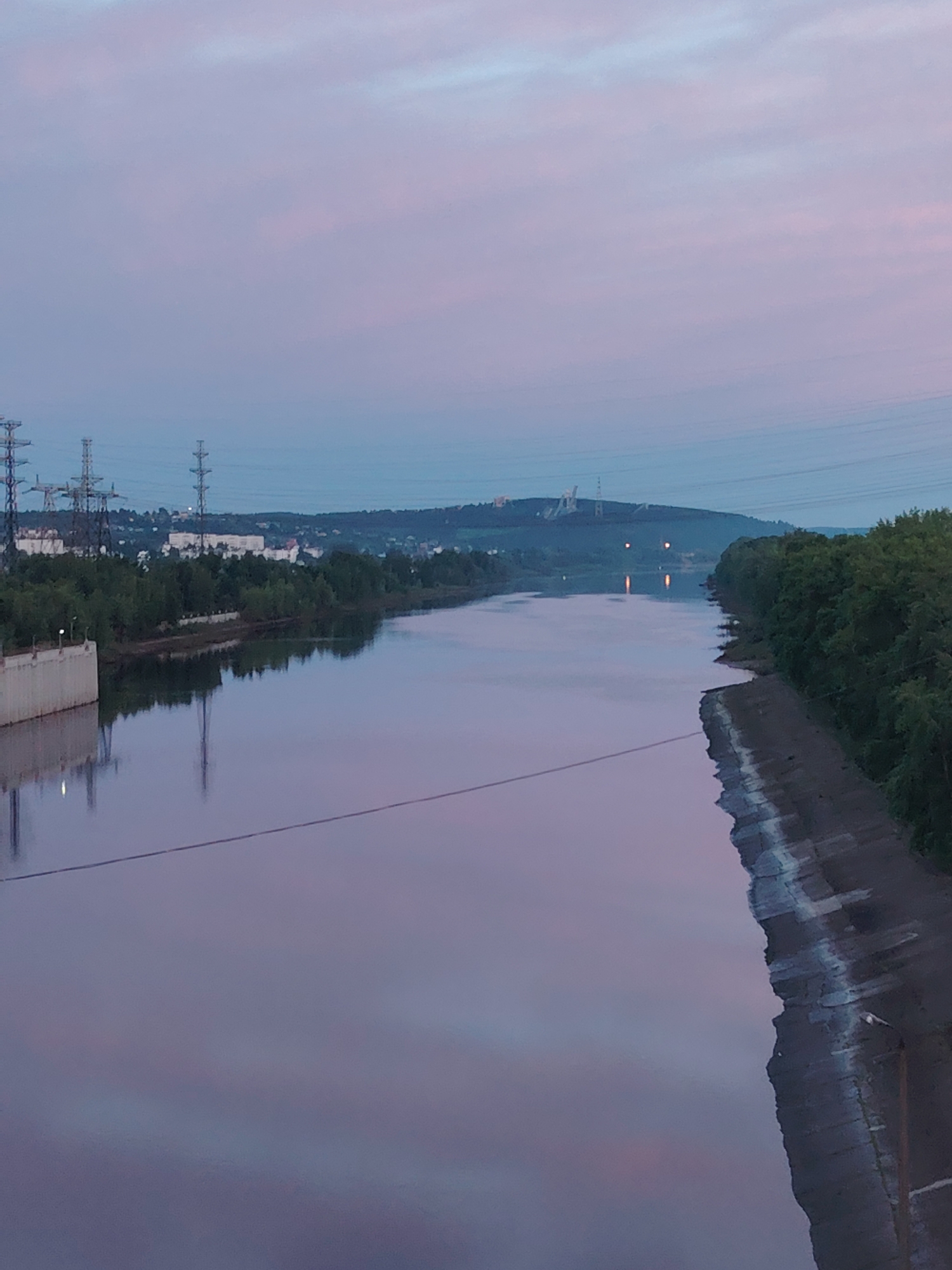 The image presents a wide river scene at either dawn or dusk, characterized by a pastel sky displaying soft pink and light blue hues beautifully reflected on the calm water surface. The left bank features a sturdy concrete retaining wall, dense green foliage, and multiple tall power transmission towers. Further behind the trees, distant urban or industrial structures are visible against a low, hilly horizon. The right bank is lined with a sloped, concrete or rip-rap embankment, also densely bordered by trees. Further down the river, artificial lights from the distant shore create shimmering vertical reflections on the water. Overhead, numerous power lines stretch horizontally across the sky. A thin, dark cable or wire is visible spanning the lower foreground, just above the water. A small, light-colored object rests on the concrete slope of the right bank near the bottom. The scene conveys a sense of peaceful, yet industrially influenced, landscape. There are no people or explicit activities taking place, and no visible text. The setting aligns with an urban riverfront environment, such as in Chaikovsky, Russia.